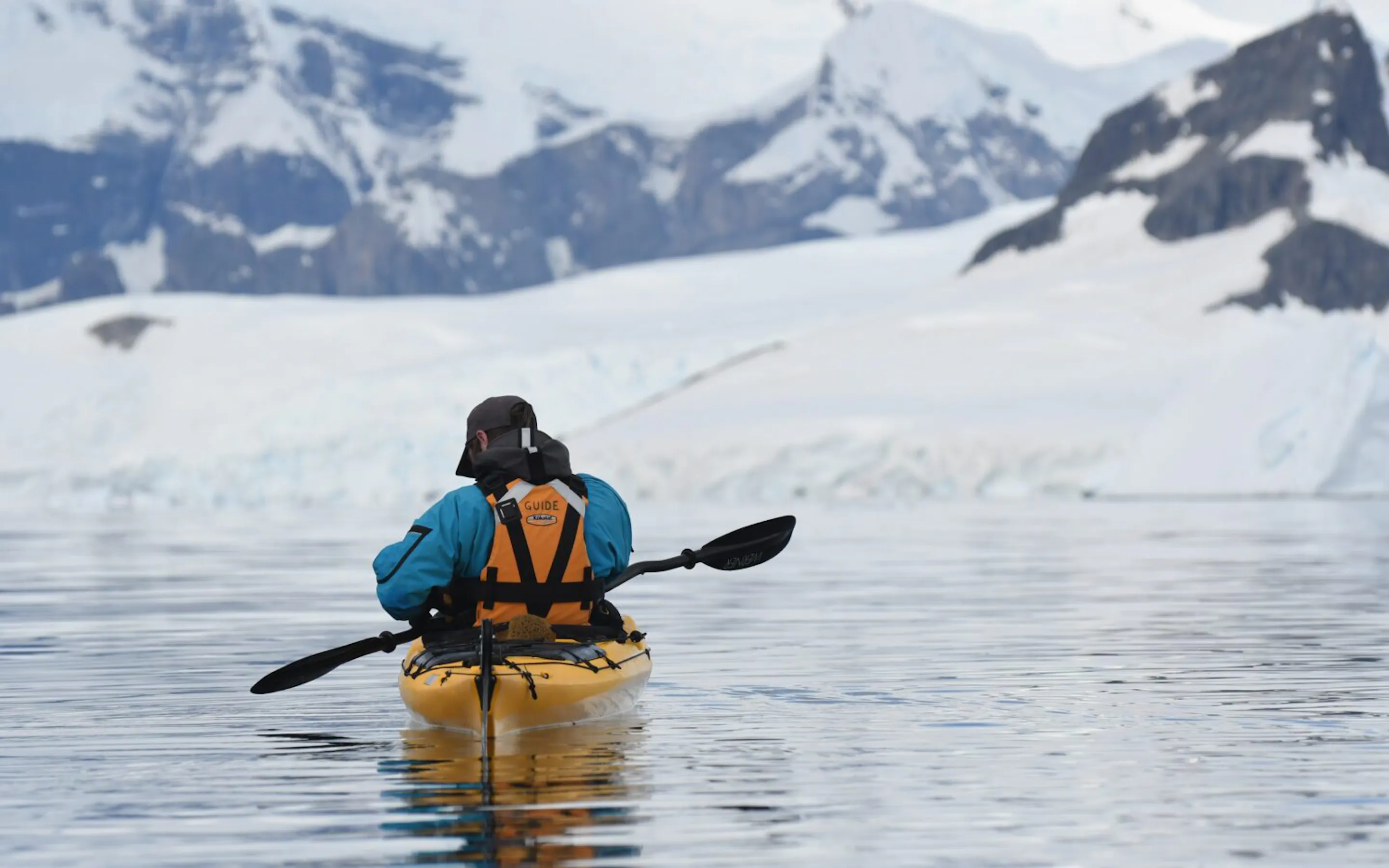 A kayaker crossing icy Antarctic waters beneath snowy peaks against the backdrop of the Antarctic Peninsula.