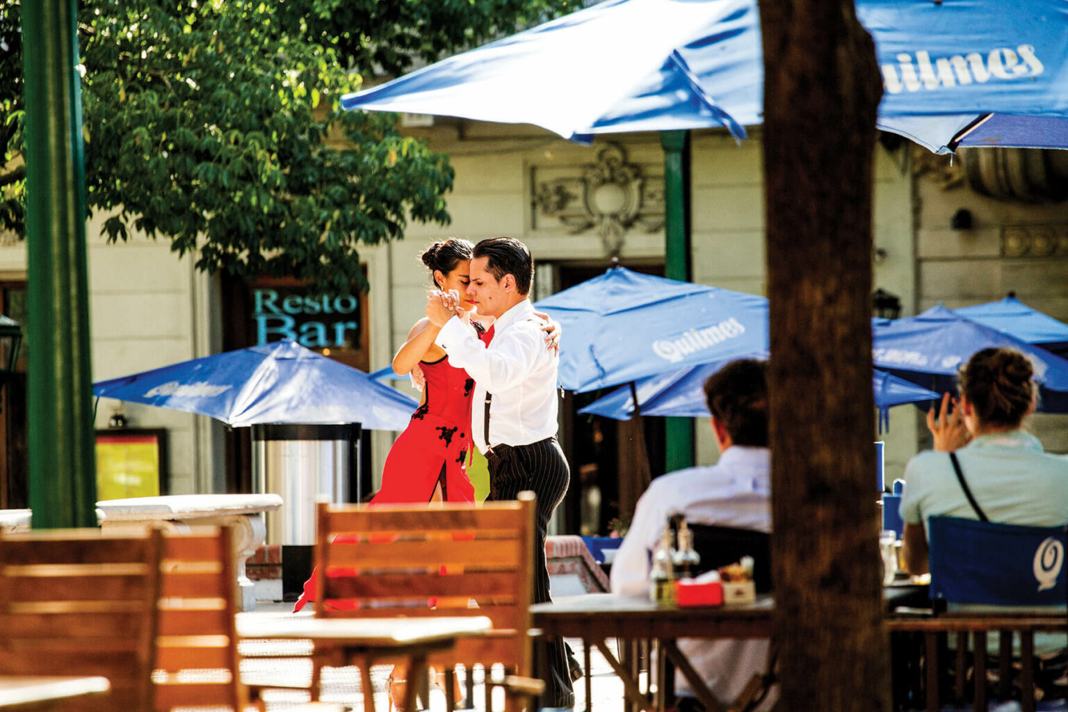 A couple dances tango beside outdoor cafe tables in Buenos Aires, with stone facades and shuttered windows behind.