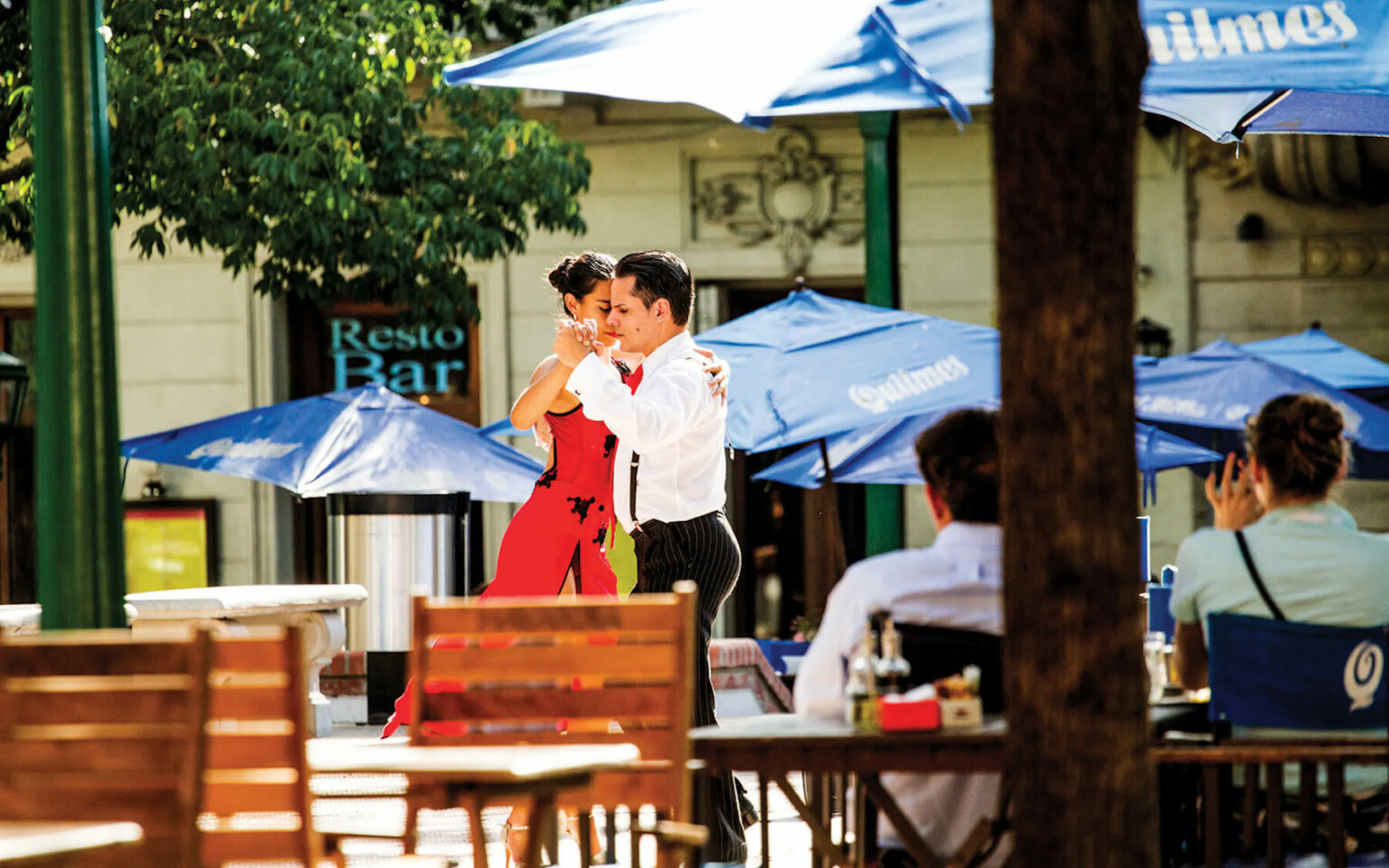 A couple dances tango beside outdoor cafe tables in Buenos Aires, with stone facades and shuttered windows behind.