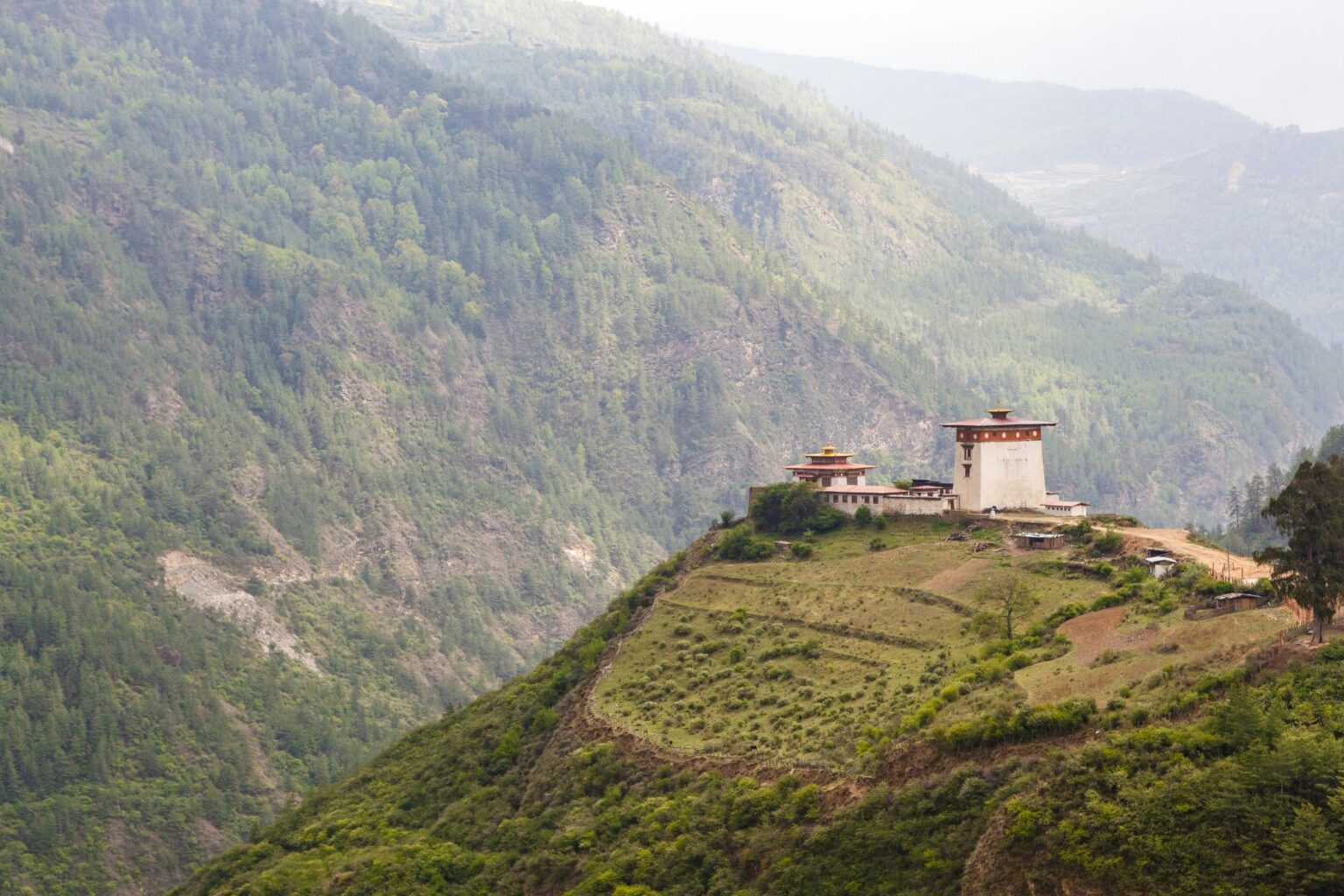 A whitewashed temple crowns a steep green hillside above terraced fields in Bhutan's remote Haa Valley under soft cloud.