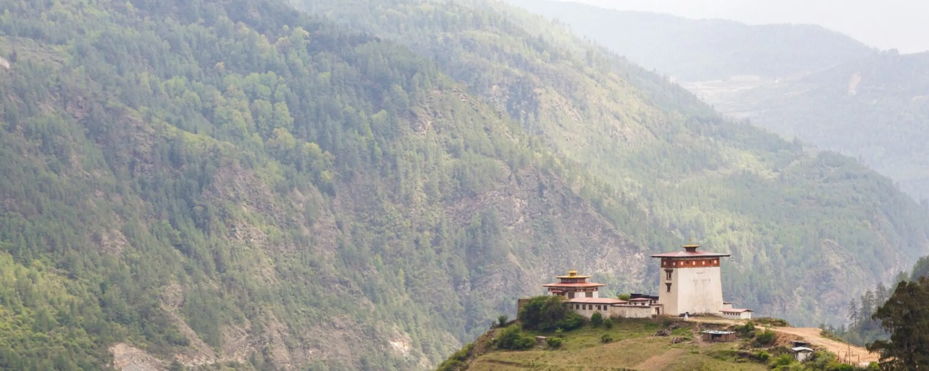 A whitewashed temple crowns a steep green hillside above terraced fields in Bhutan's remote Haa Valley under soft cloud.