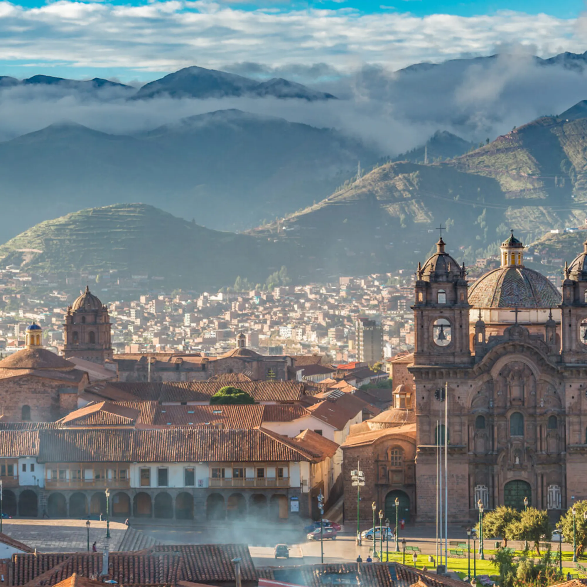 Cusco rooftops and cathedral towers rise beneath layered mountains in Peru, with tiled buildings filling the foreground.