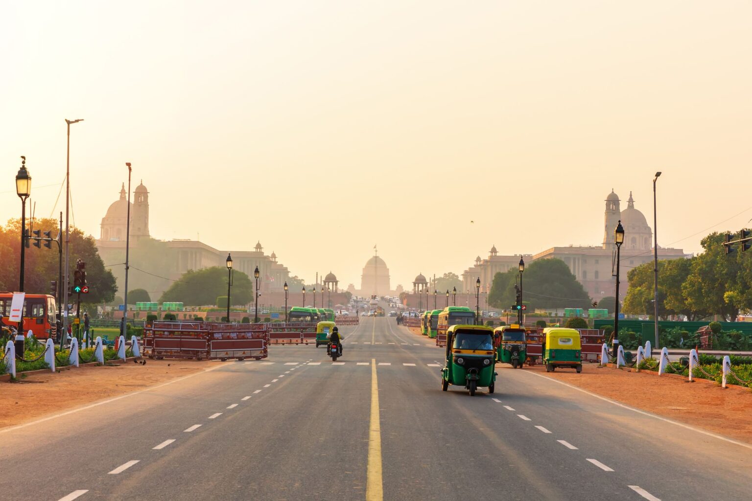 A view down a broad Delhi boulevard, where rickshaws and motorbikes move through soft haze.