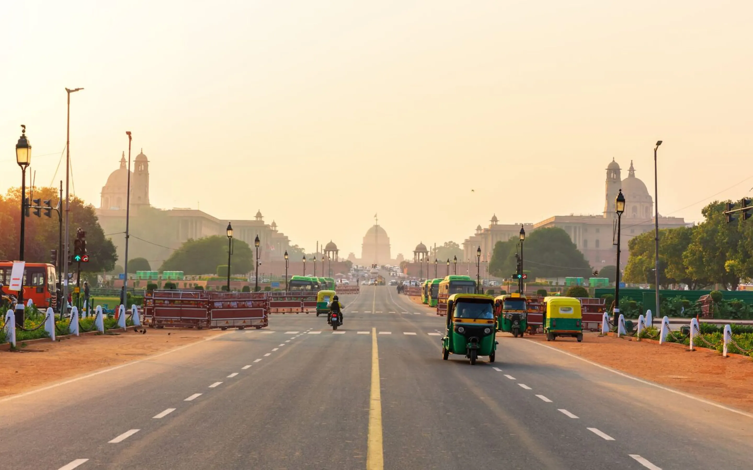 A view down a broad Delhi boulevard, where rickshaws and motorbikes move through soft haze.
