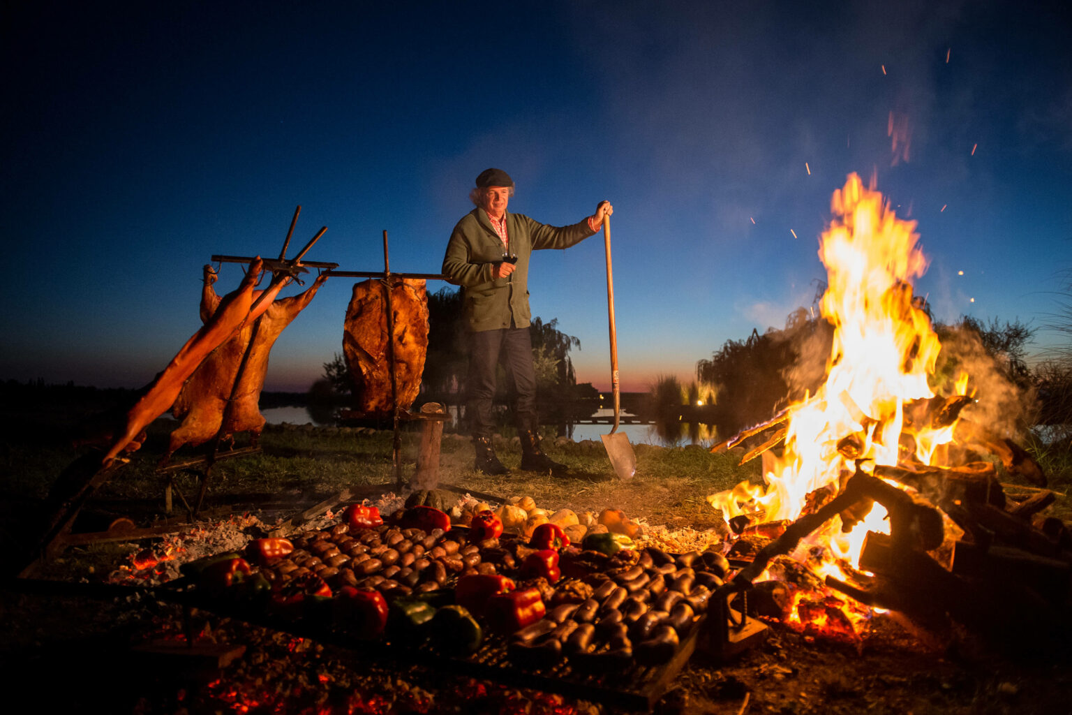 A man tends an open fire beside vineyard rows in Mendoza, Argentina, with low vines and dusk sky behind him.
