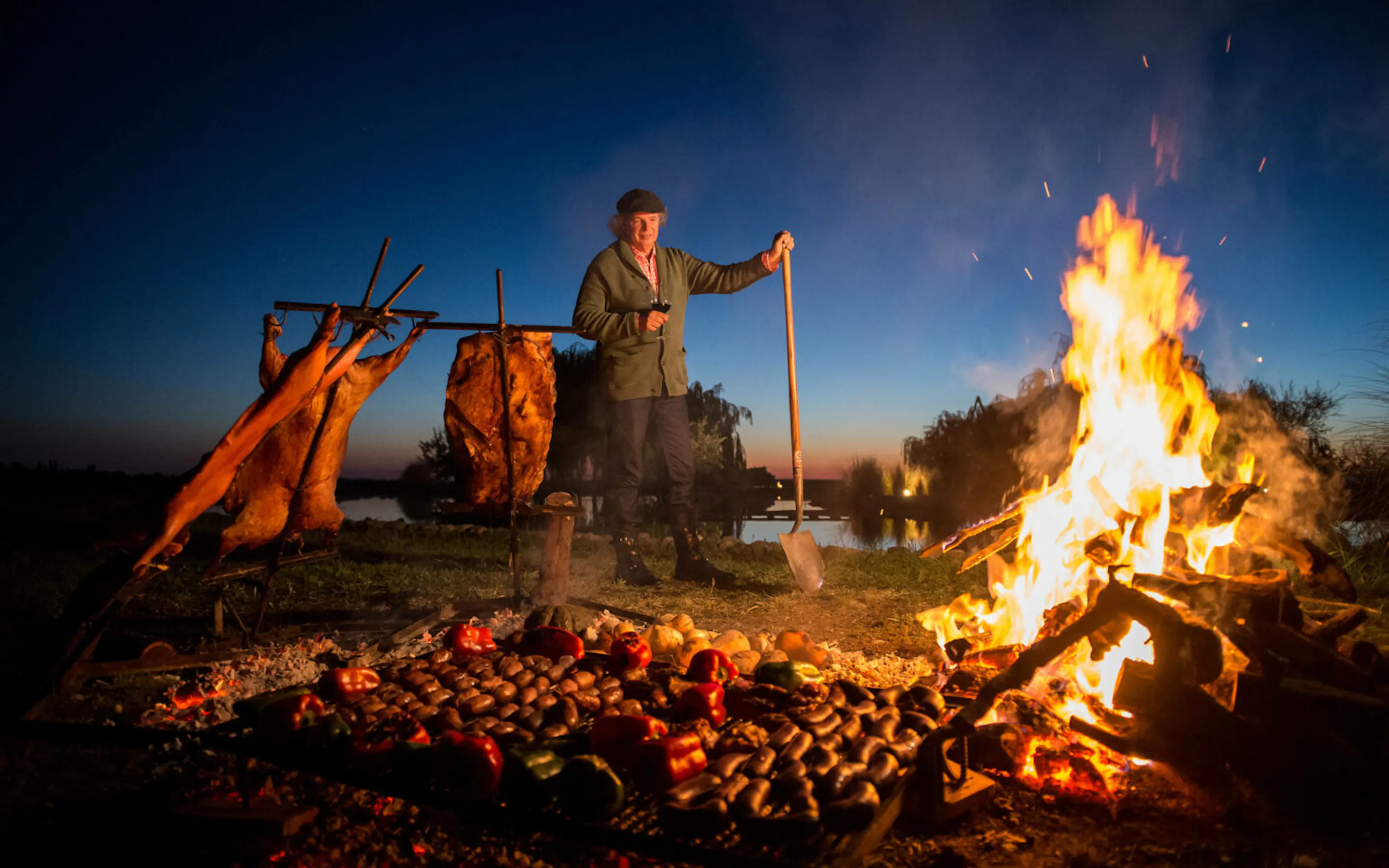 A man tends an open fire beside vineyard rows in Mendoza, Argentina, with low vines and dusk sky behind him.