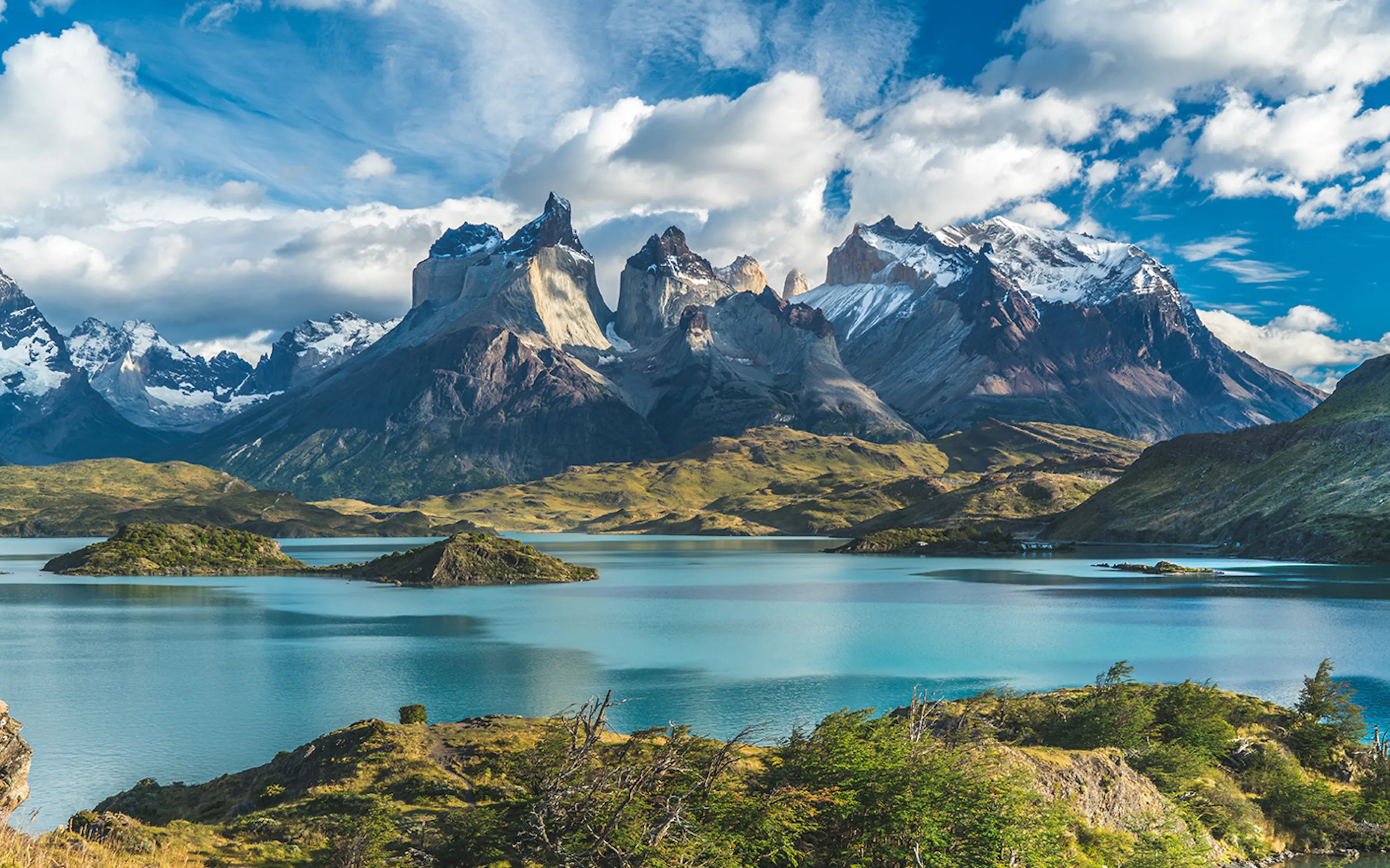 Jagged Torres del Paine peaks rise above a blue lake, with rocky headlands and bright Patagonian light across the water.