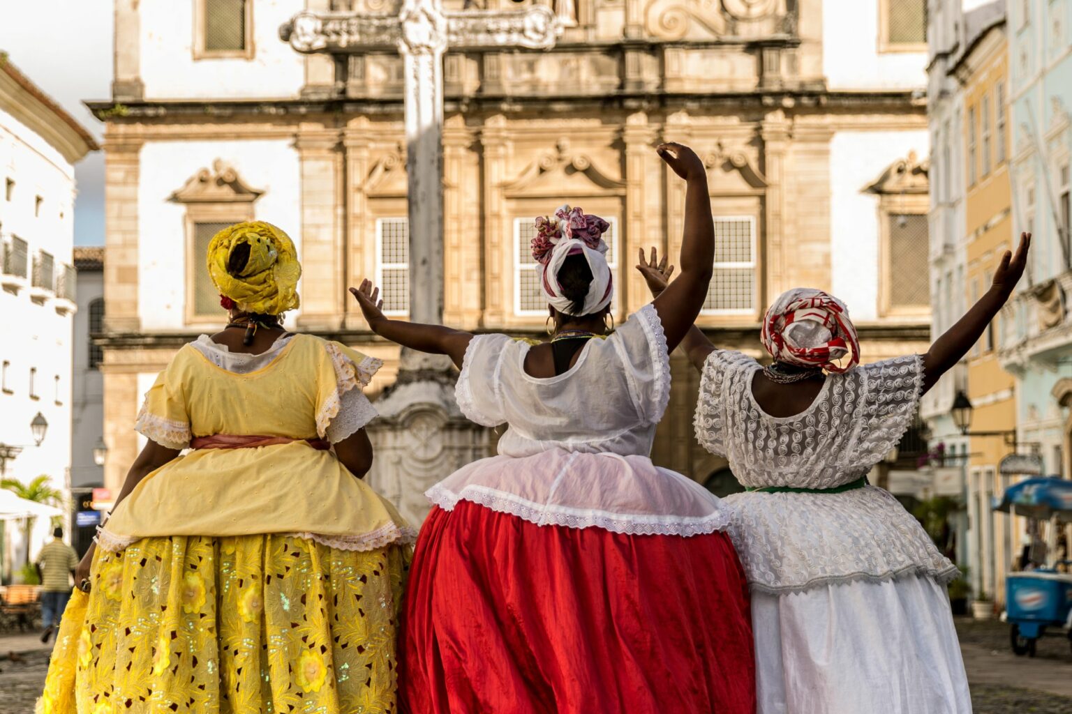 Three women in bright dresses and headwraps walk beside a church square in Bahia, framed by colonial facades.