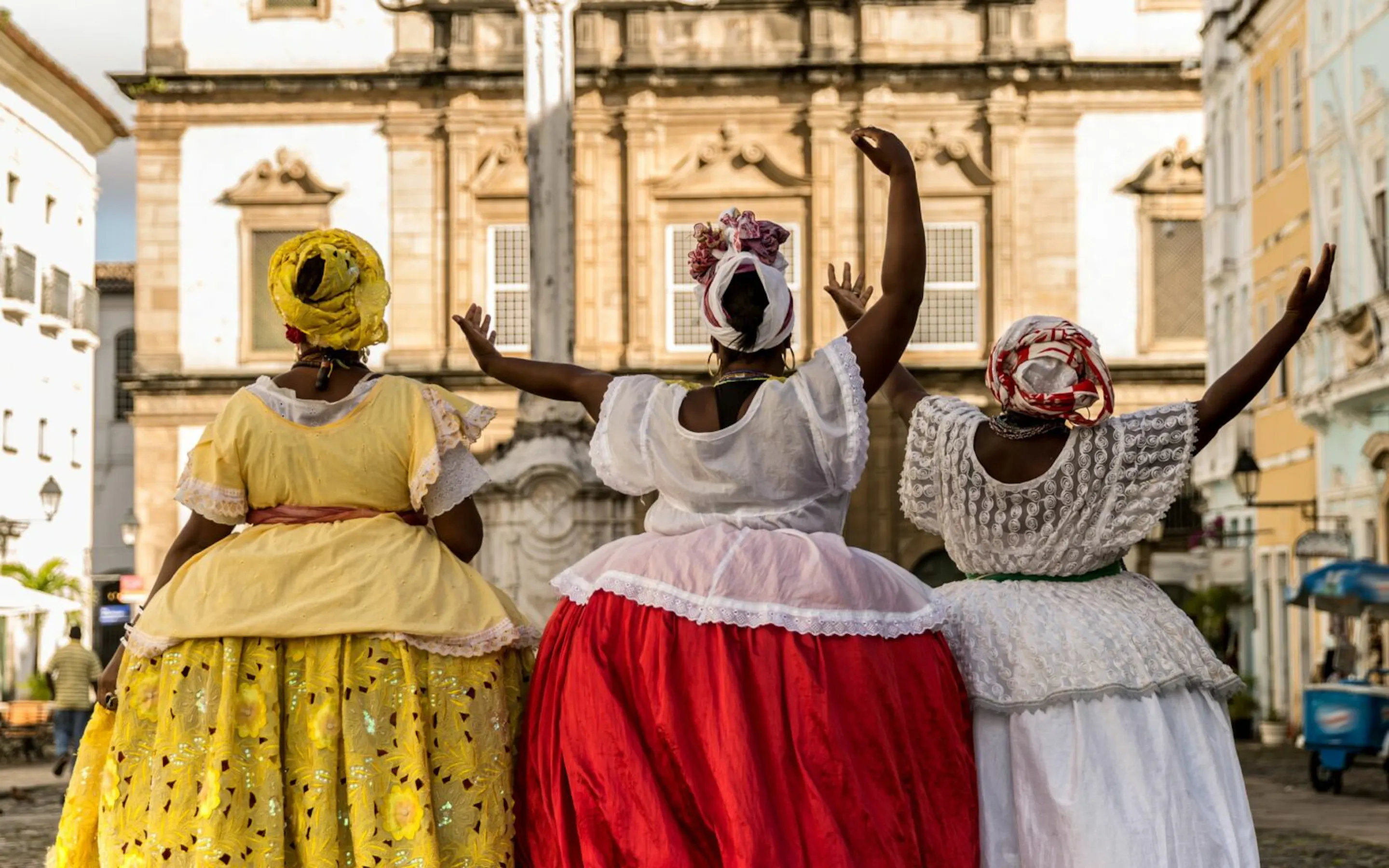 Three women in bright dresses and headwraps walk beside a church square in Bahia, framed by colonial facades.