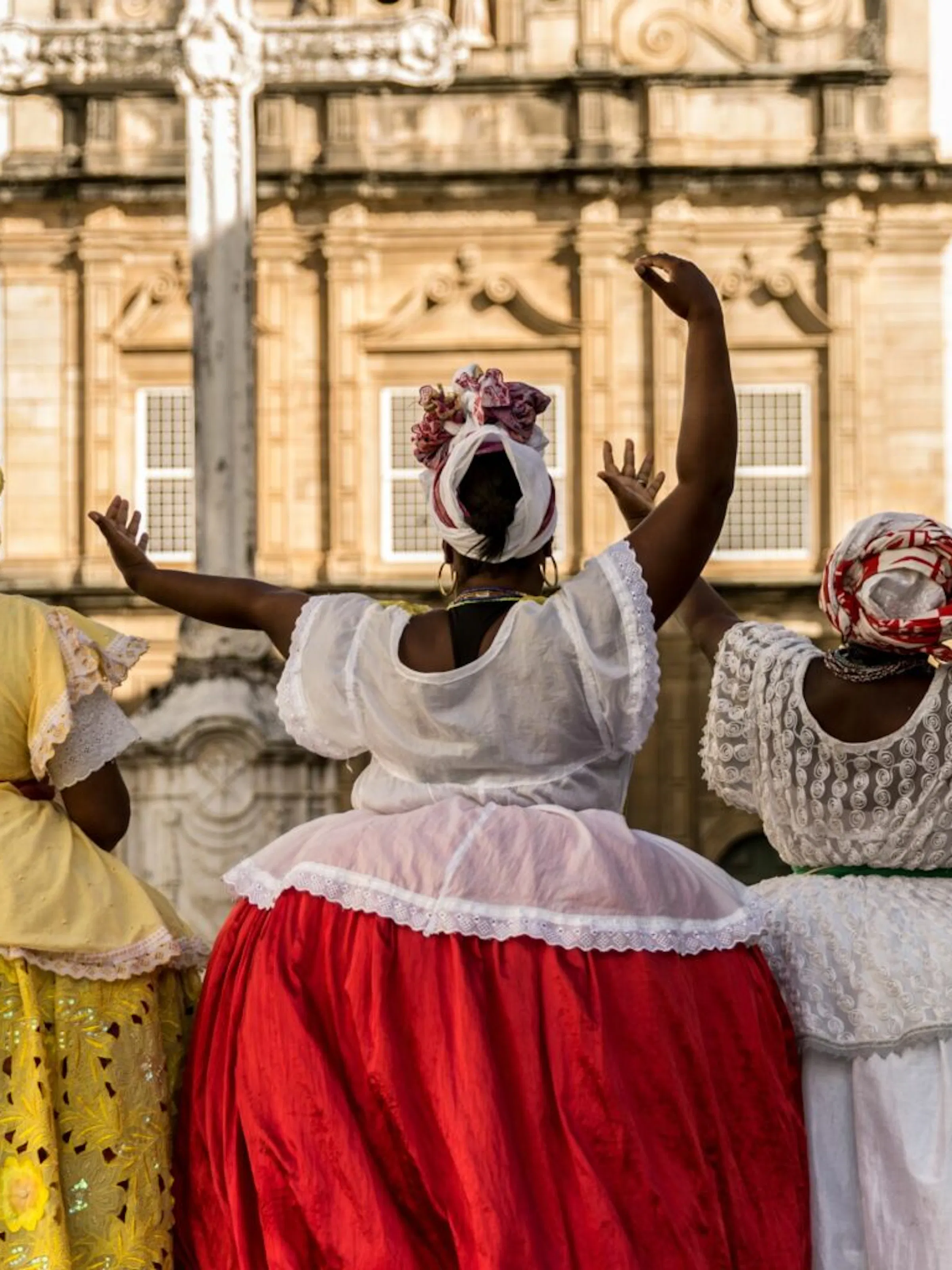 Three women in bright dresses and headwraps walk beside a church square in Bahia, framed by colonial facades.
