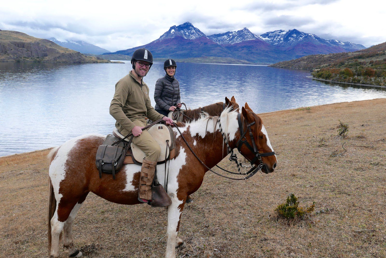 Two riders on horseback travel beside a Patagonian lake at Explora, with distant peaks rising beyond the shore.