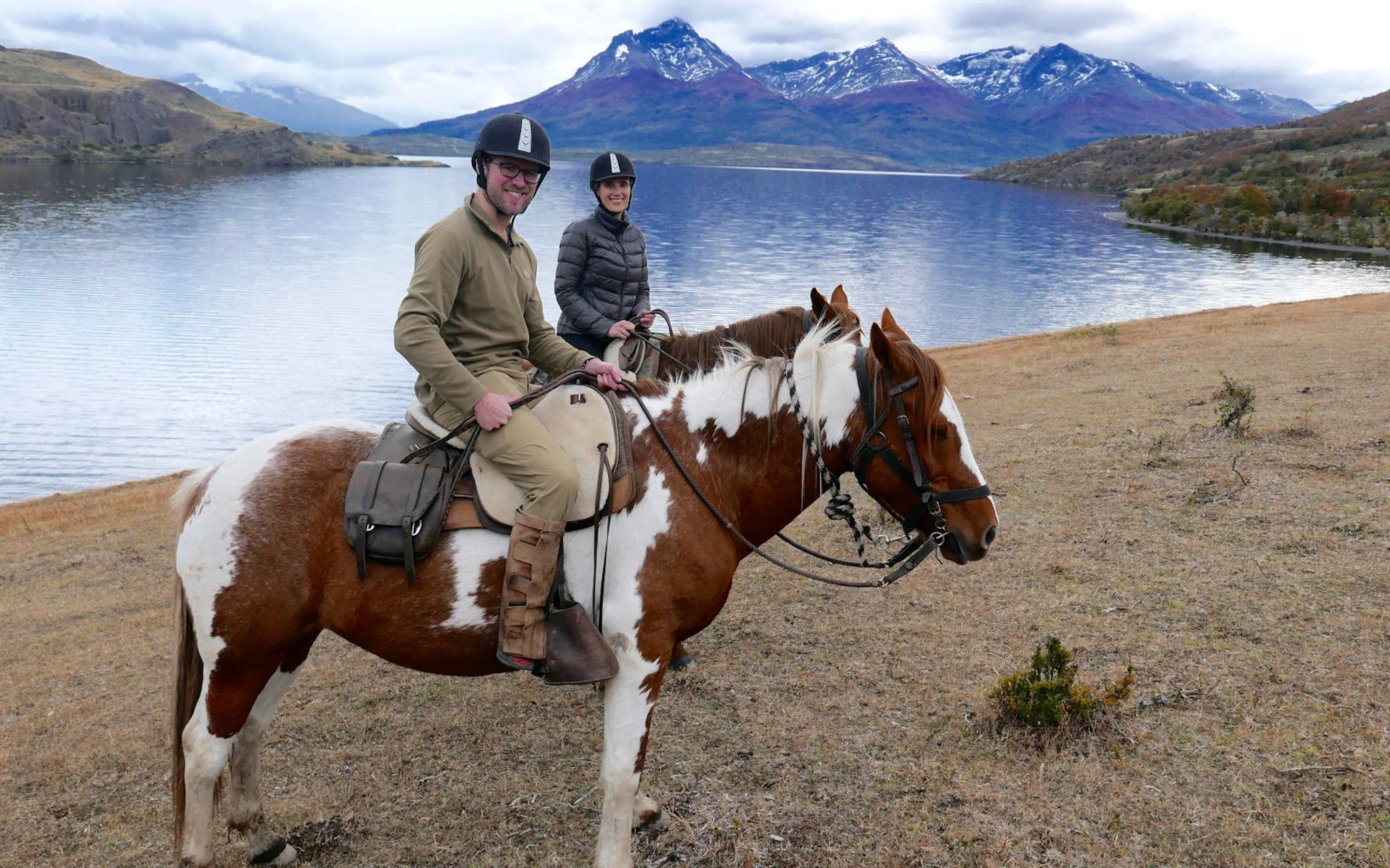 Two riders on horseback travel beside a Patagonian lake at Explora, with distant peaks rising beyond the shore.