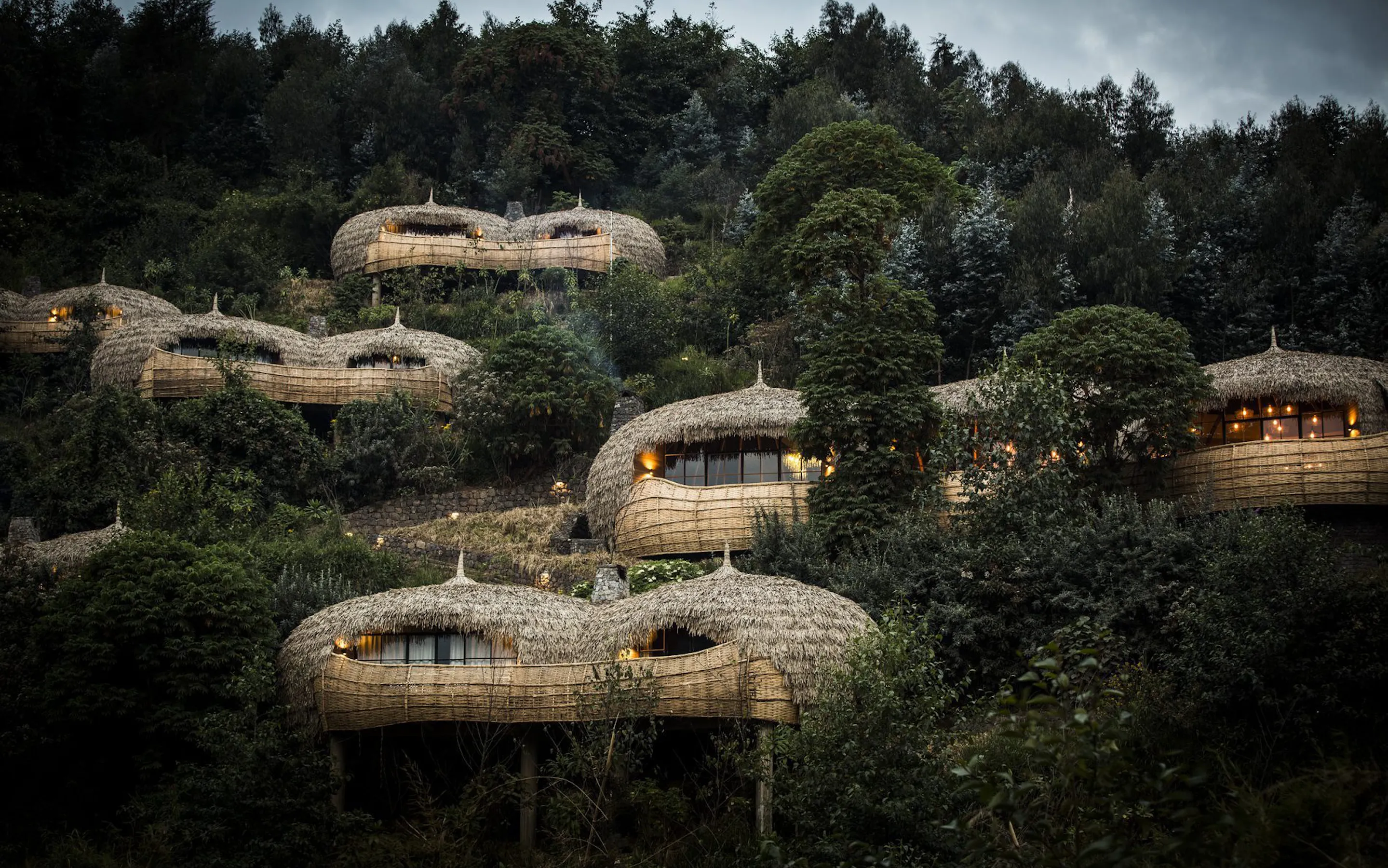 Thatched villas curve into a lush hillside at Bisate Lodge, overlooking forest, gardens, and distant Rwanda peaks.