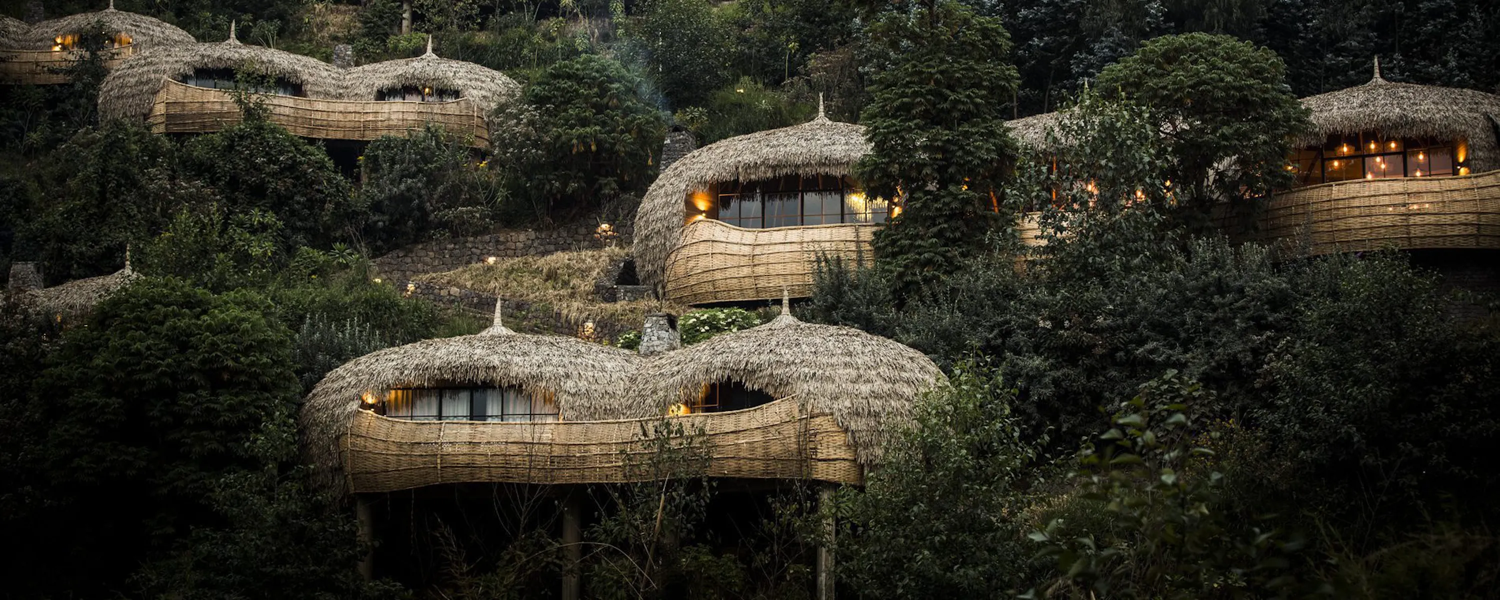 Thatched villas curve into a lush hillside at Bisate Lodge, overlooking forest, gardens, and distant Rwanda peaks.