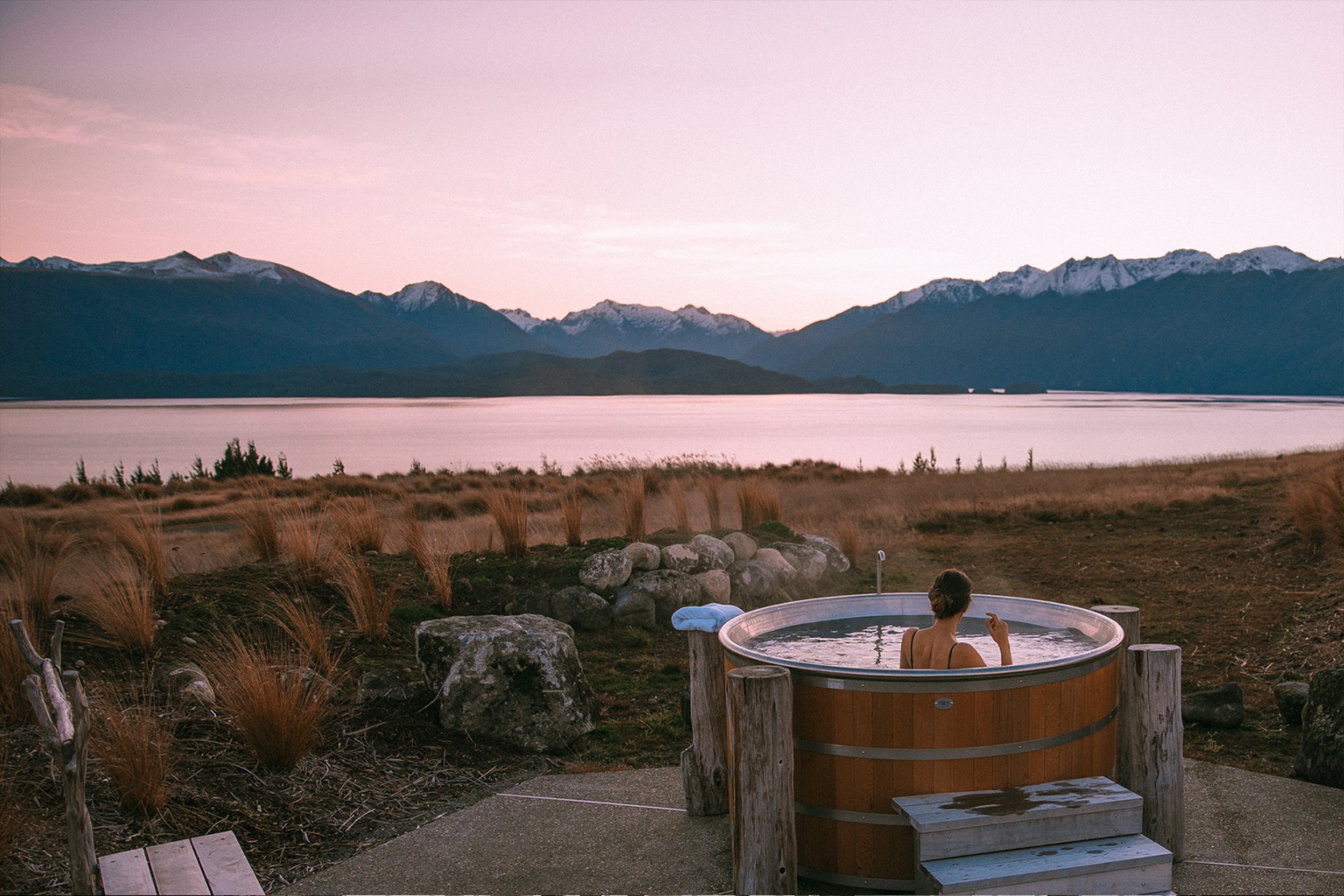 A woman relaxes in a cedar hot tub at Fiordland Lodge, with lake water and layered hills stretching beyond.