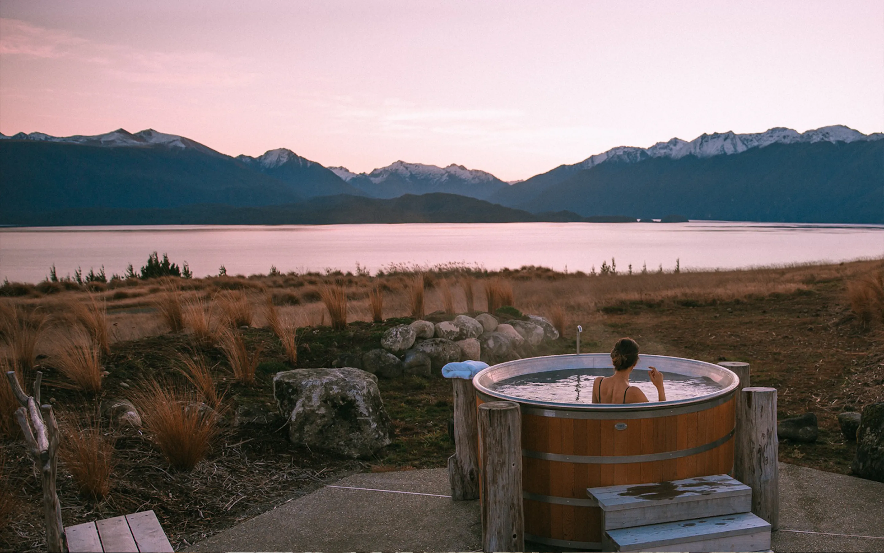 A woman relaxes in a cedar hot tub at Fiordland Lodge, with lake water and layered hills stretching beyond.