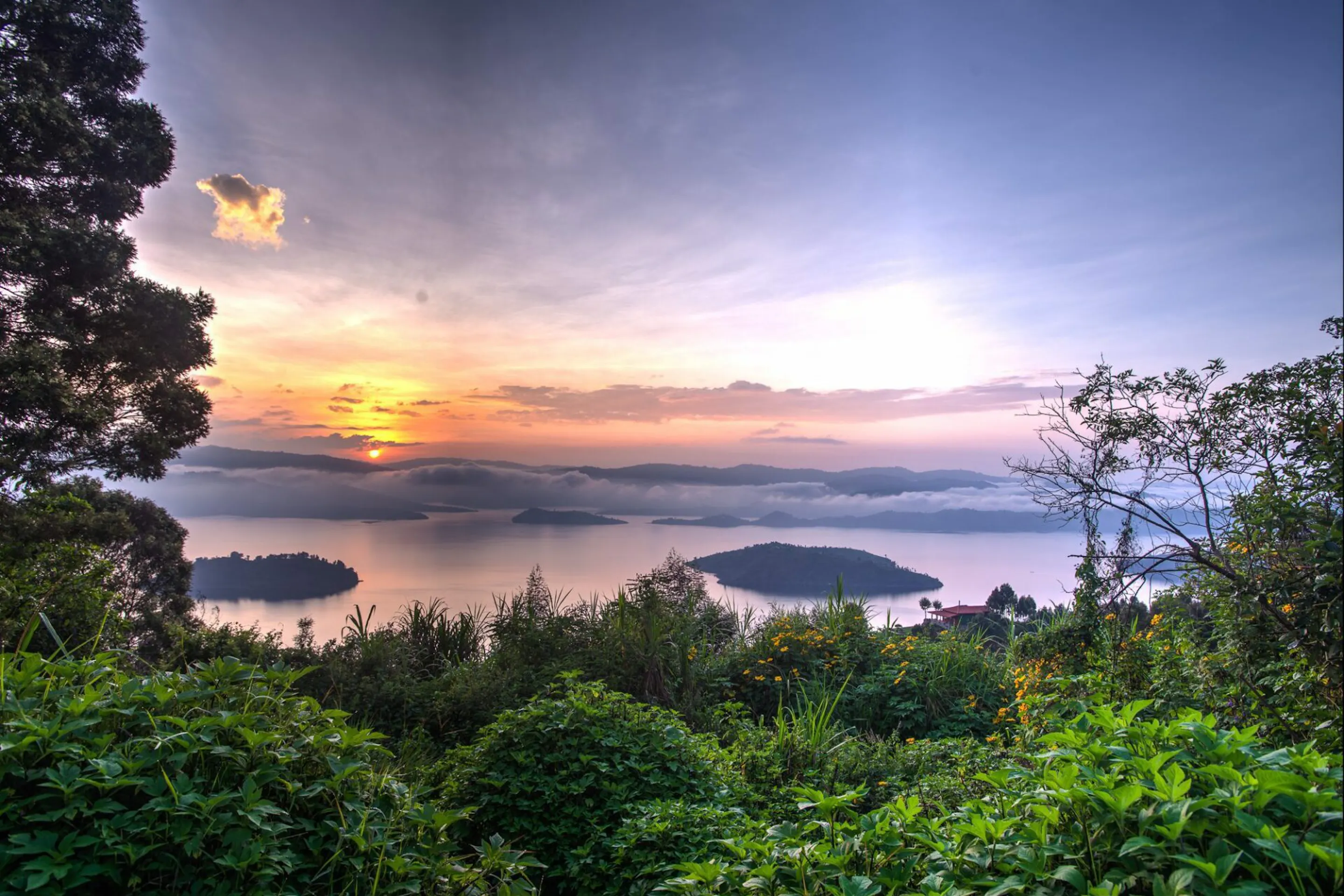 Soft pink light settles over the lakes below Virunga Lodge, framed by leafy trees and distant Rwanda hills.