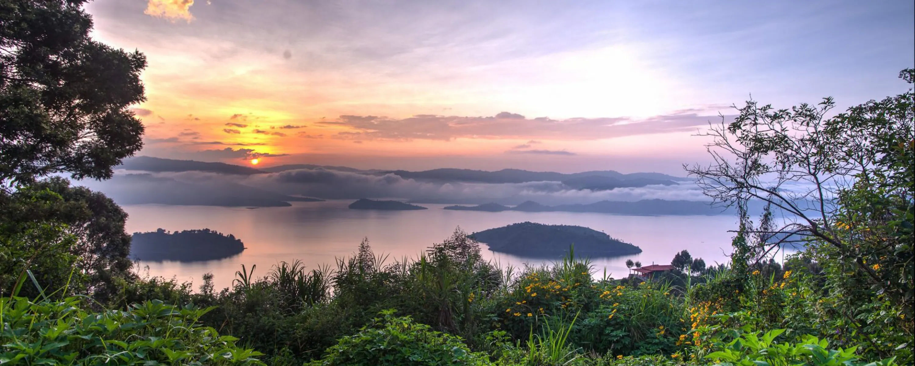 Soft pink light settles over the lakes below Virunga Lodge, framed by leafy trees and distant Rwanda hills.