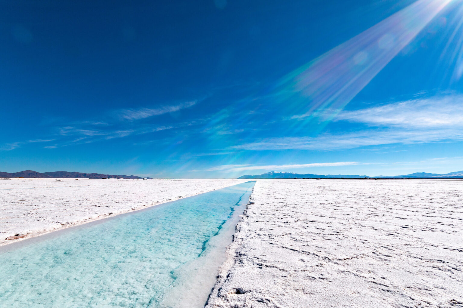 A straight channel cuts through a bright salt flat in Salta, Argentina, beneath a clear blue sky and distant peaks.