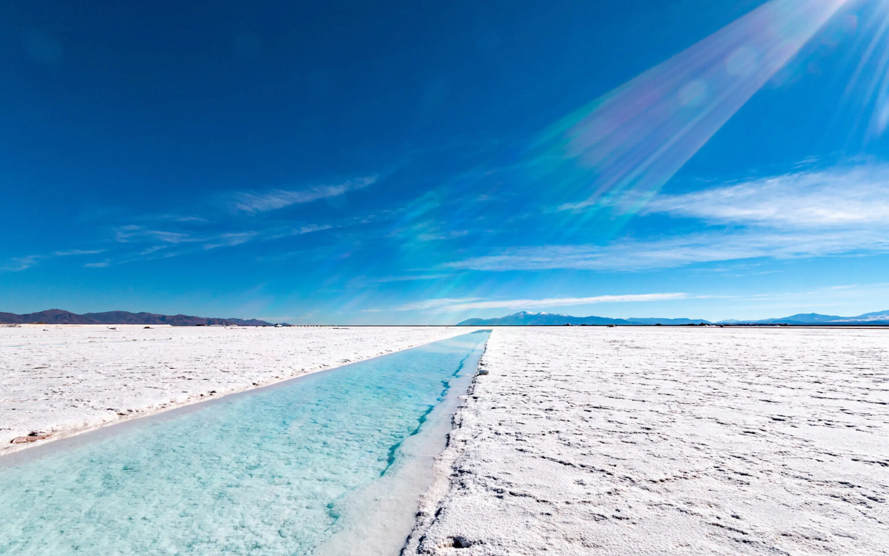 A straight channel cuts through a bright salt flat in Salta, Argentina, beneath a clear blue sky and distant peaks.