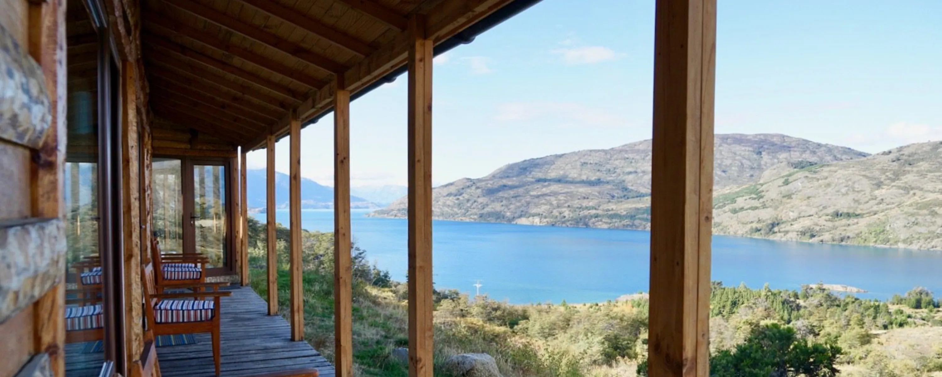 Wooden chairs line a covered porch at Mallin Colorado, overlooking Lake General Carrera and pale hills beyond.