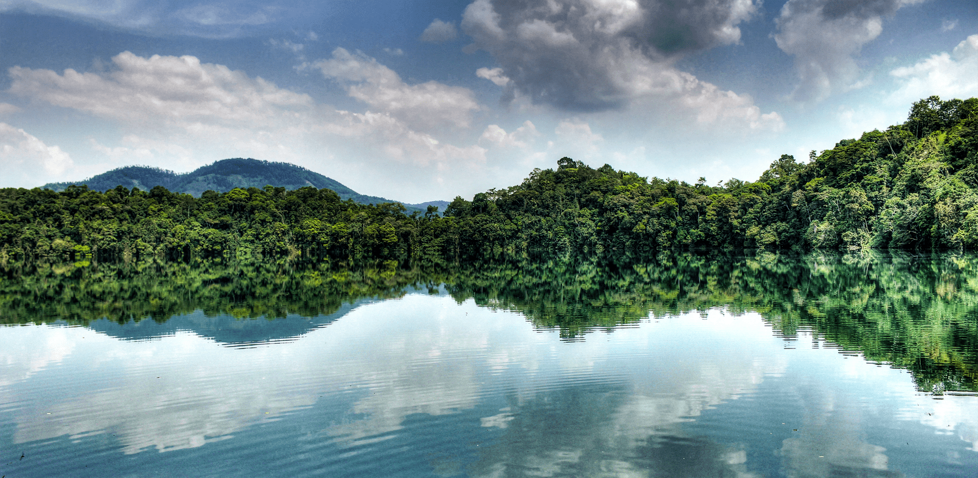 A crater lake reflects forested hills and cloudy sky in Uganda's Kibale Forest, with green slopes at the water's edge.
