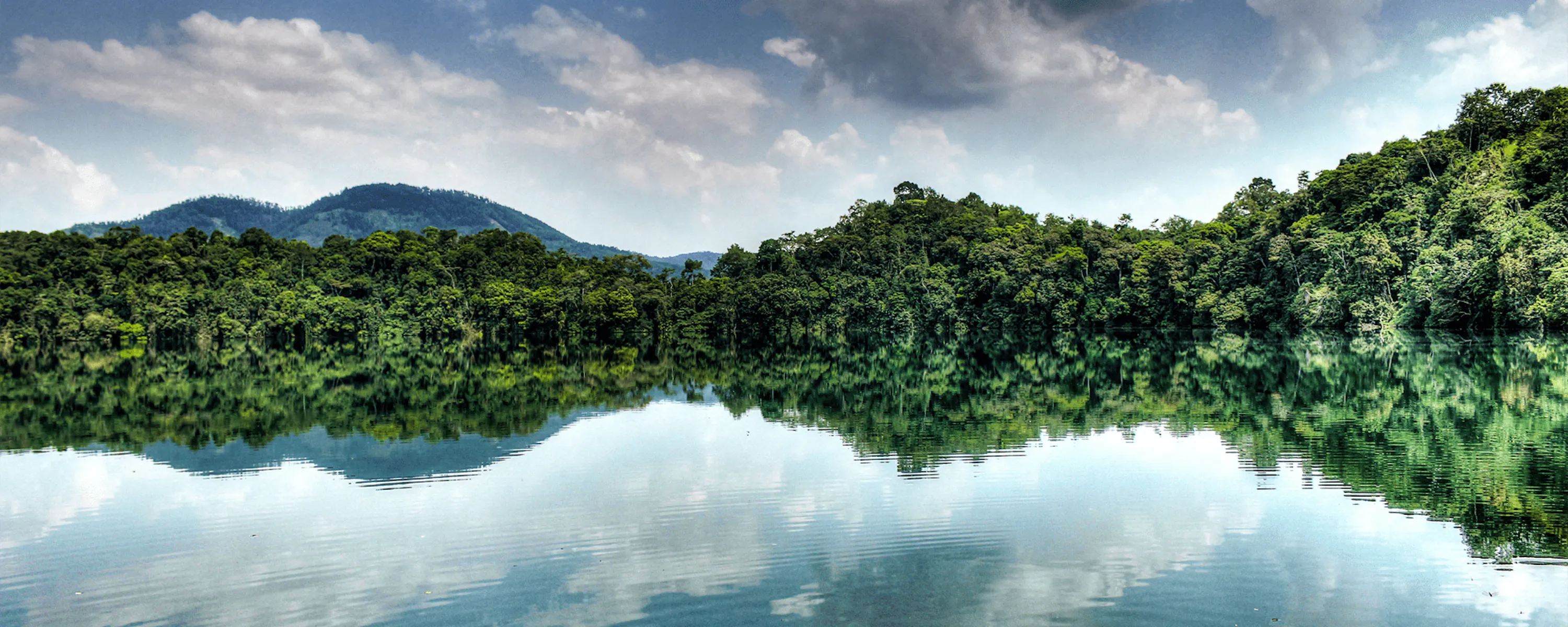 A crater lake reflects forested hills and cloudy sky in Uganda's Kibale Forest, with green slopes at the water's edge.