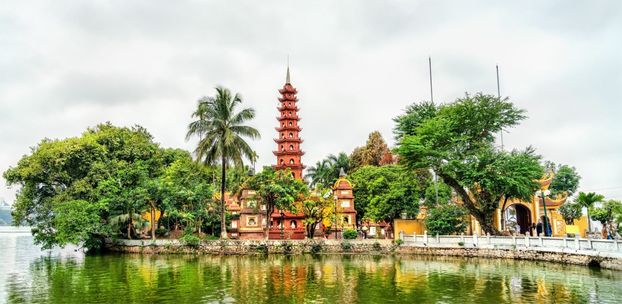 A red pagoda rises beside a calm lake, framed by palms and trees with soft reflections in the water below.