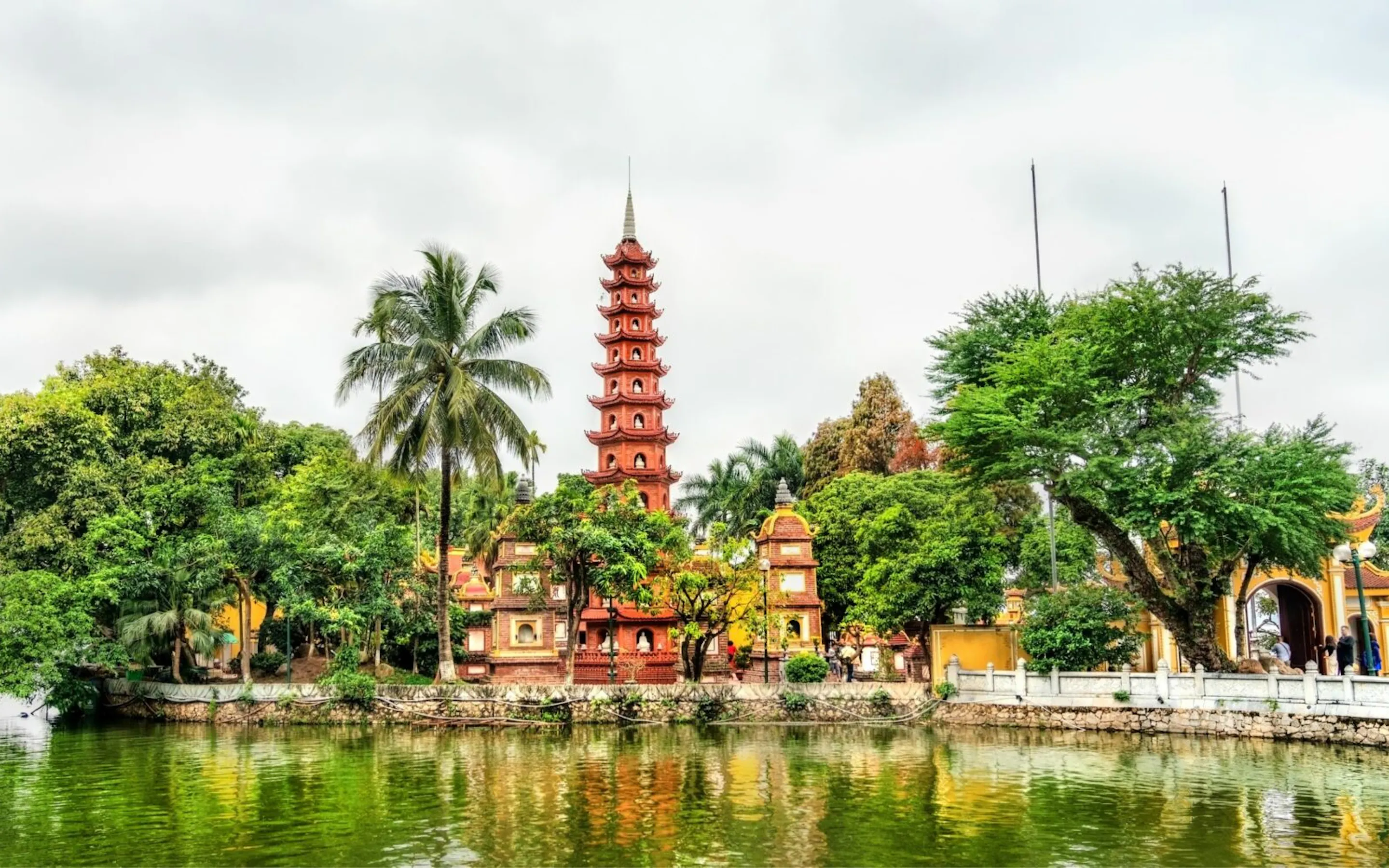 A red pagoda rises beside a calm lake, framed by palms and trees with soft reflections in the water below.