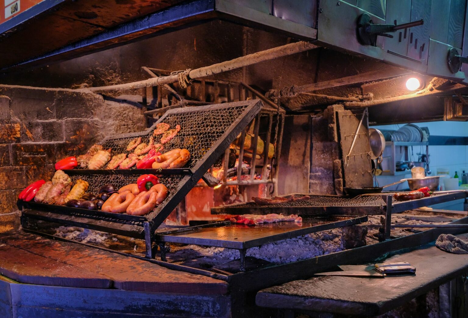 Cuts of meat roast over open flames at Montevideo's port market, with glowing coals and steel grills below.