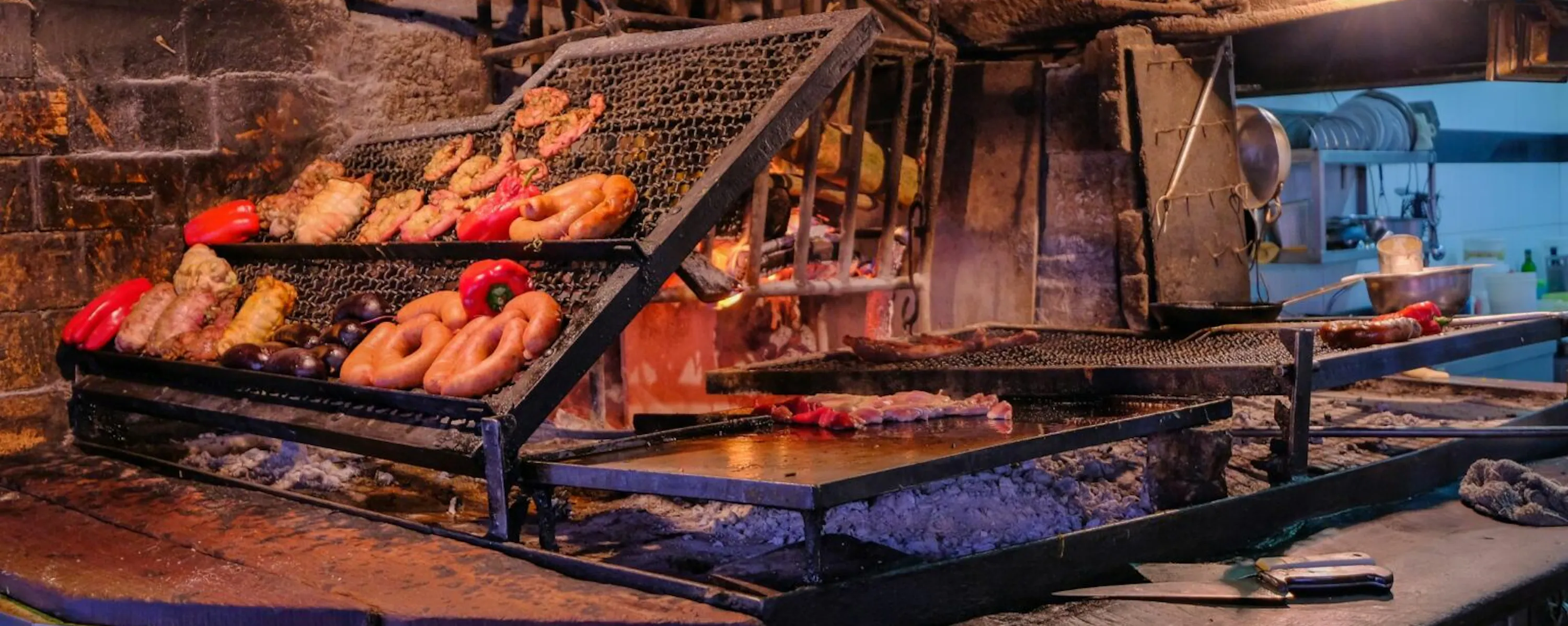 Cuts of meat roast over open flames at Montevideo's port market, with glowing coals and steel grills below.