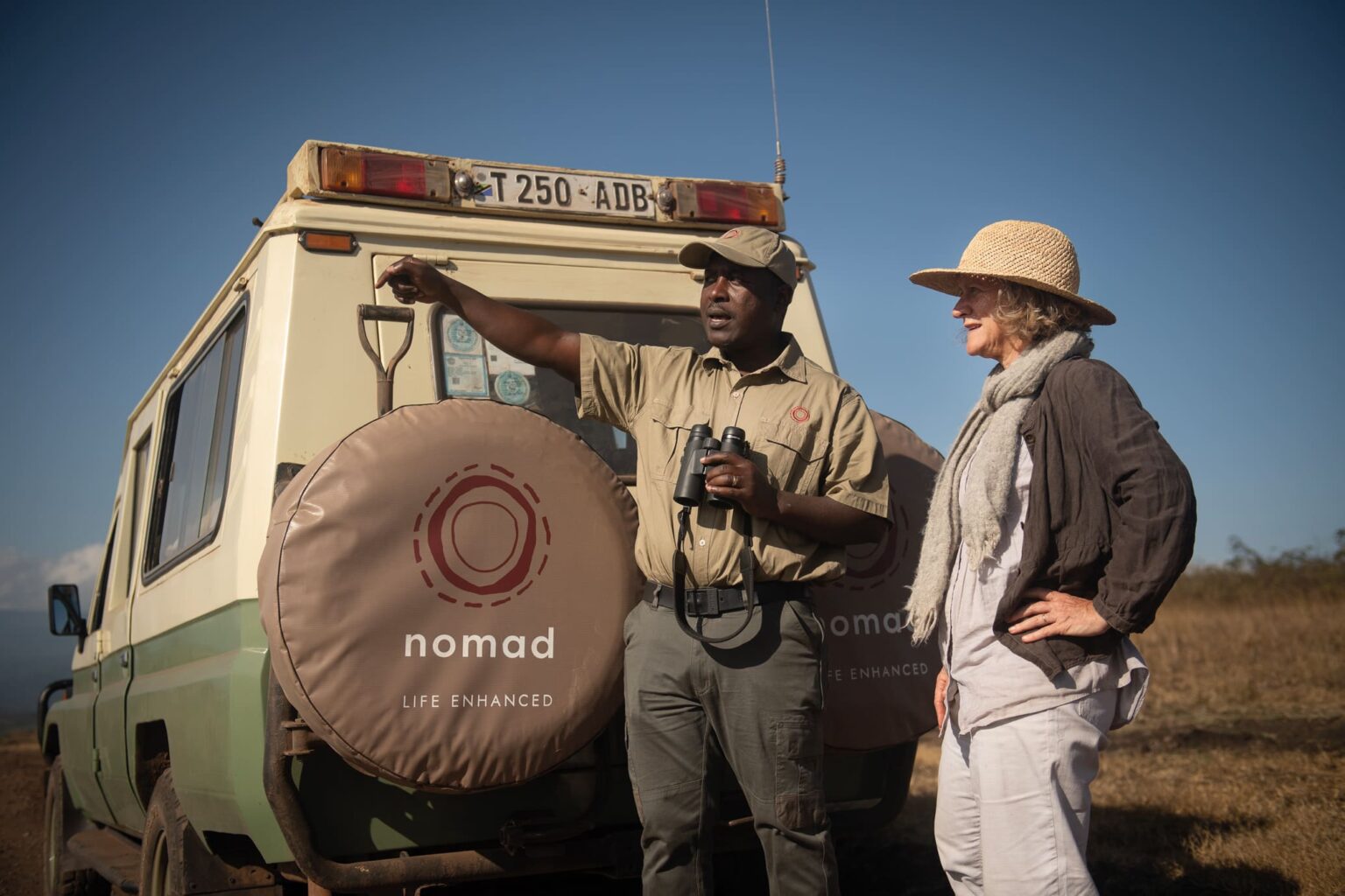 A smiling Nomad guide stands beside a safari vehicle in Tanzania, binoculars in hand, with guest waiting nearby.
