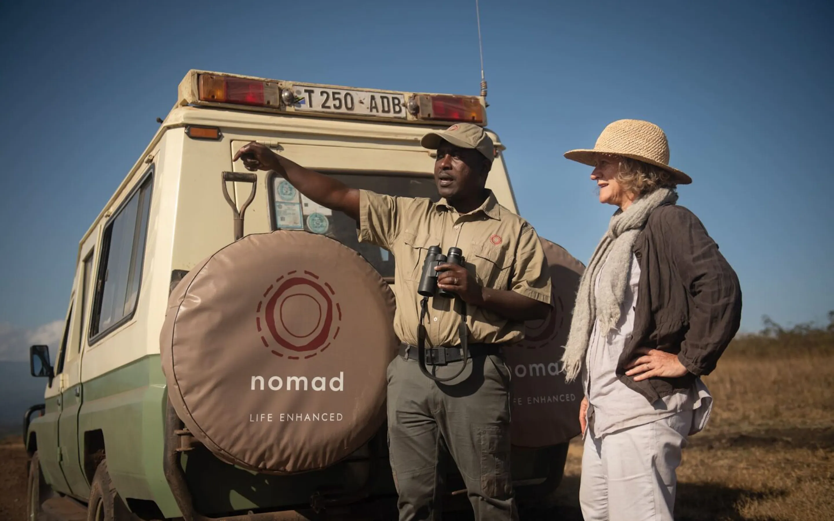 A smiling Nomad guide stands beside a safari vehicle in Tanzania, binoculars in hand, with guest waiting nearby.