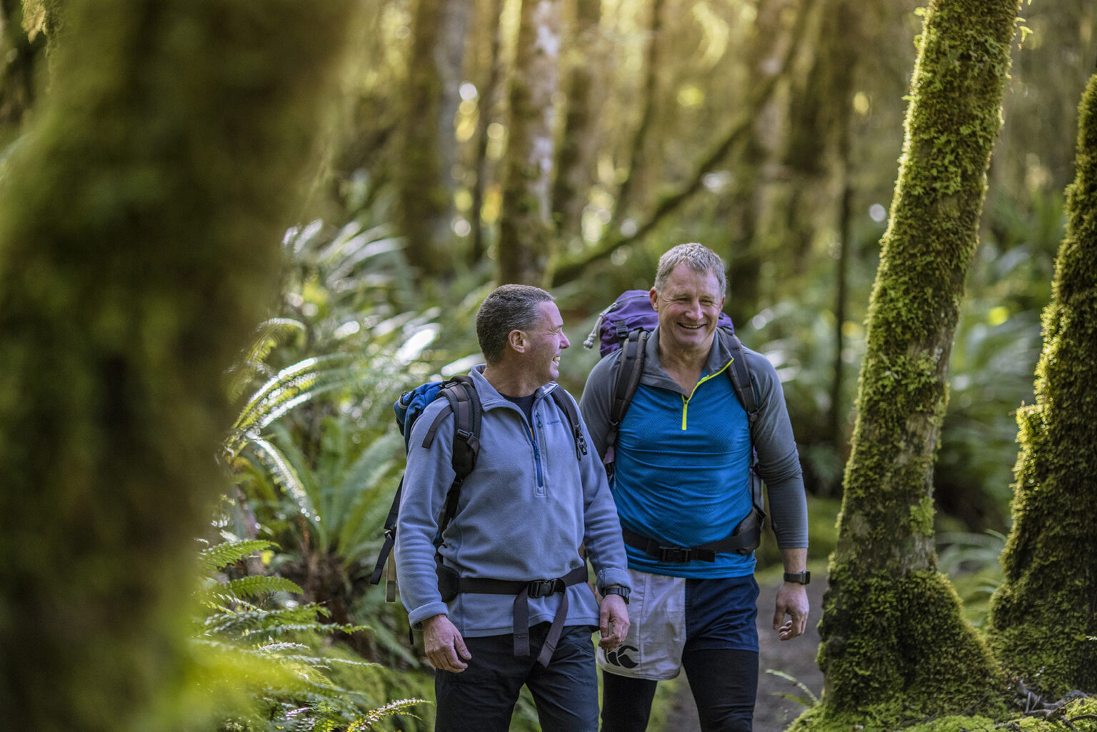 Two hikers walking through a mossy forest on the Kepler Track against the backdrop of New Zealand's Fiordland.