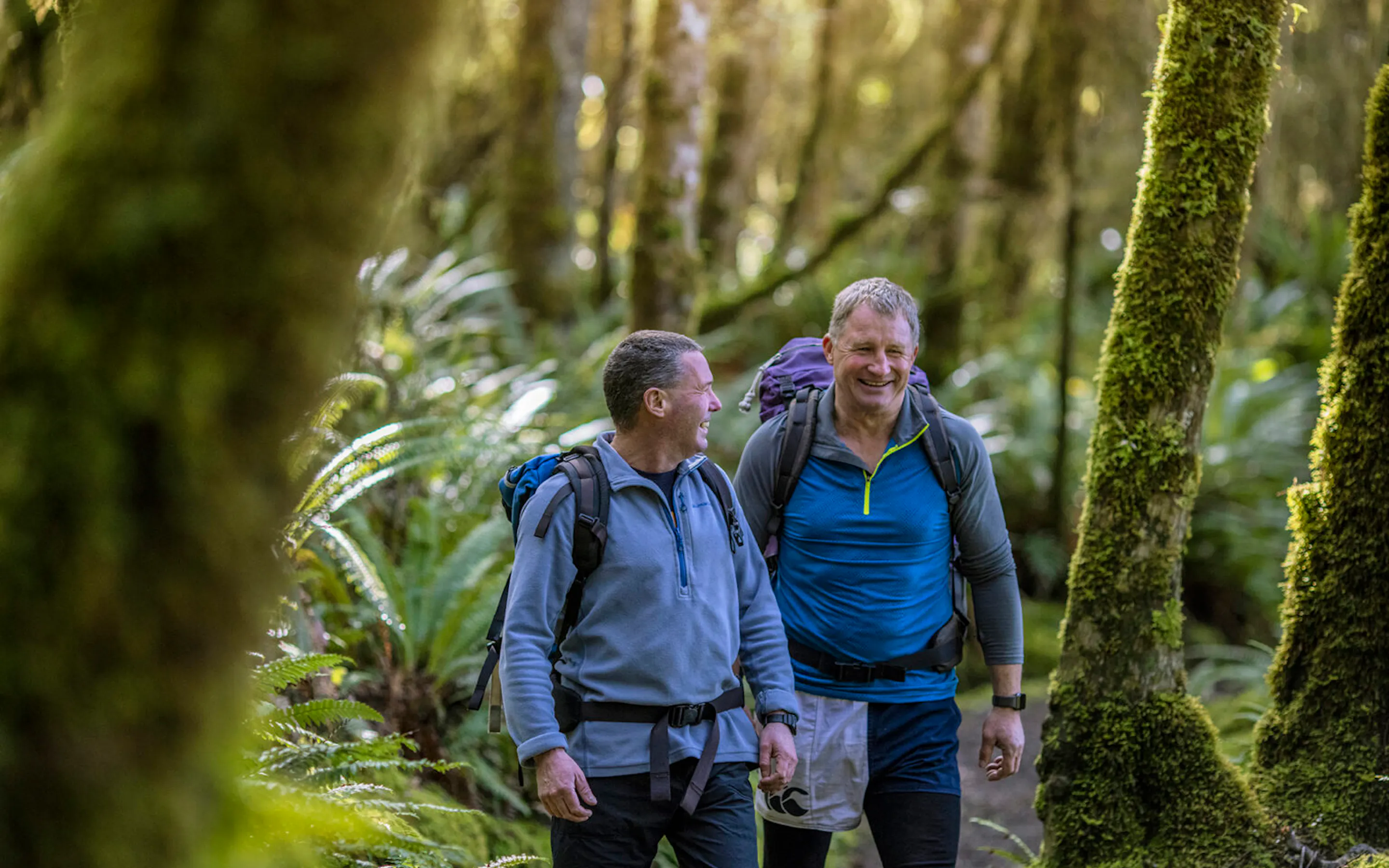 Two hikers walking through a mossy forest on the Kepler Track against the backdrop of New Zealand's Fiordland.