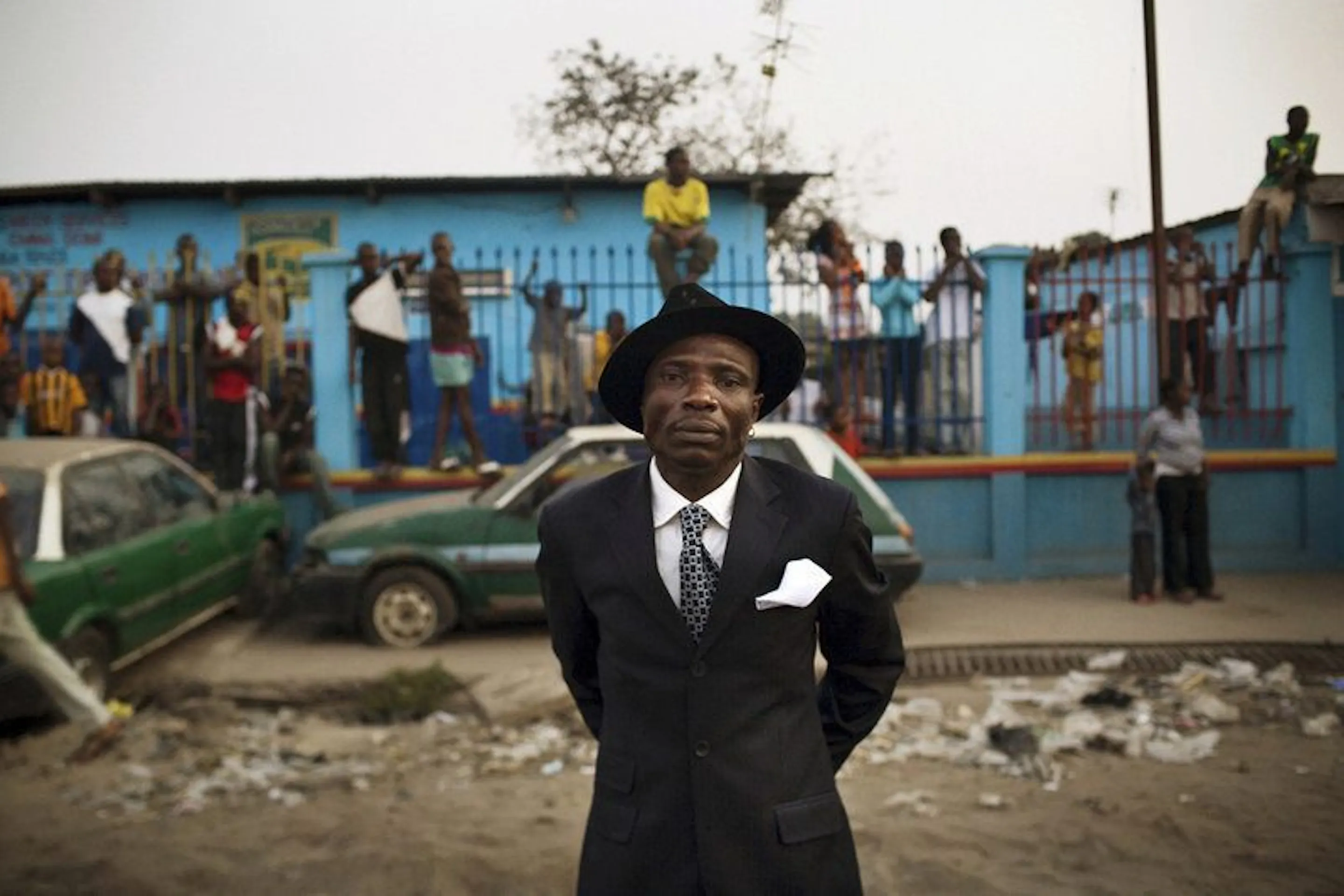 A sharply dressed sapeur stands before a lively Brazzaville street scene, framed by friends and bright color.