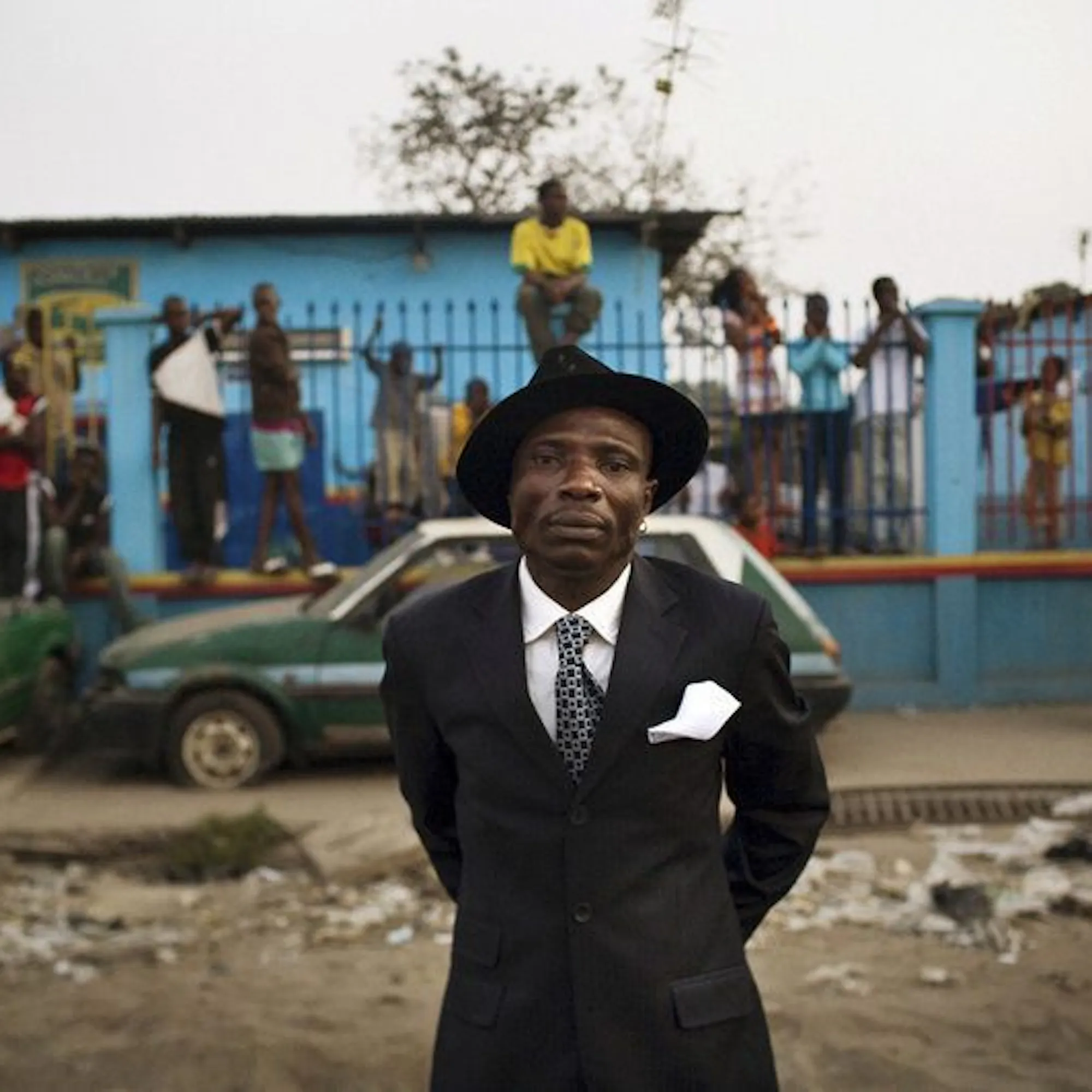 A sharply dressed sapeur stands before a lively Brazzaville street scene, framed by friends and bright color.