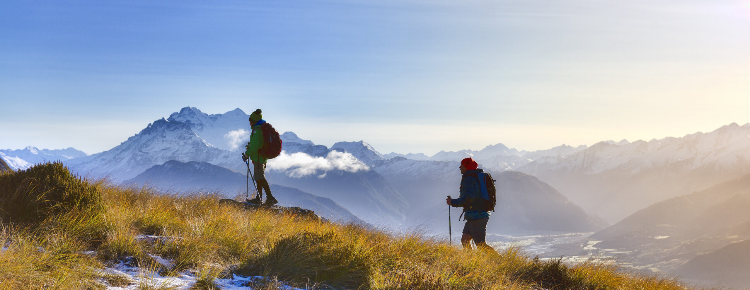 Two hikers stand on a ridge above Fiordland's misty mountains, with layered peaks fading into low cloud below.