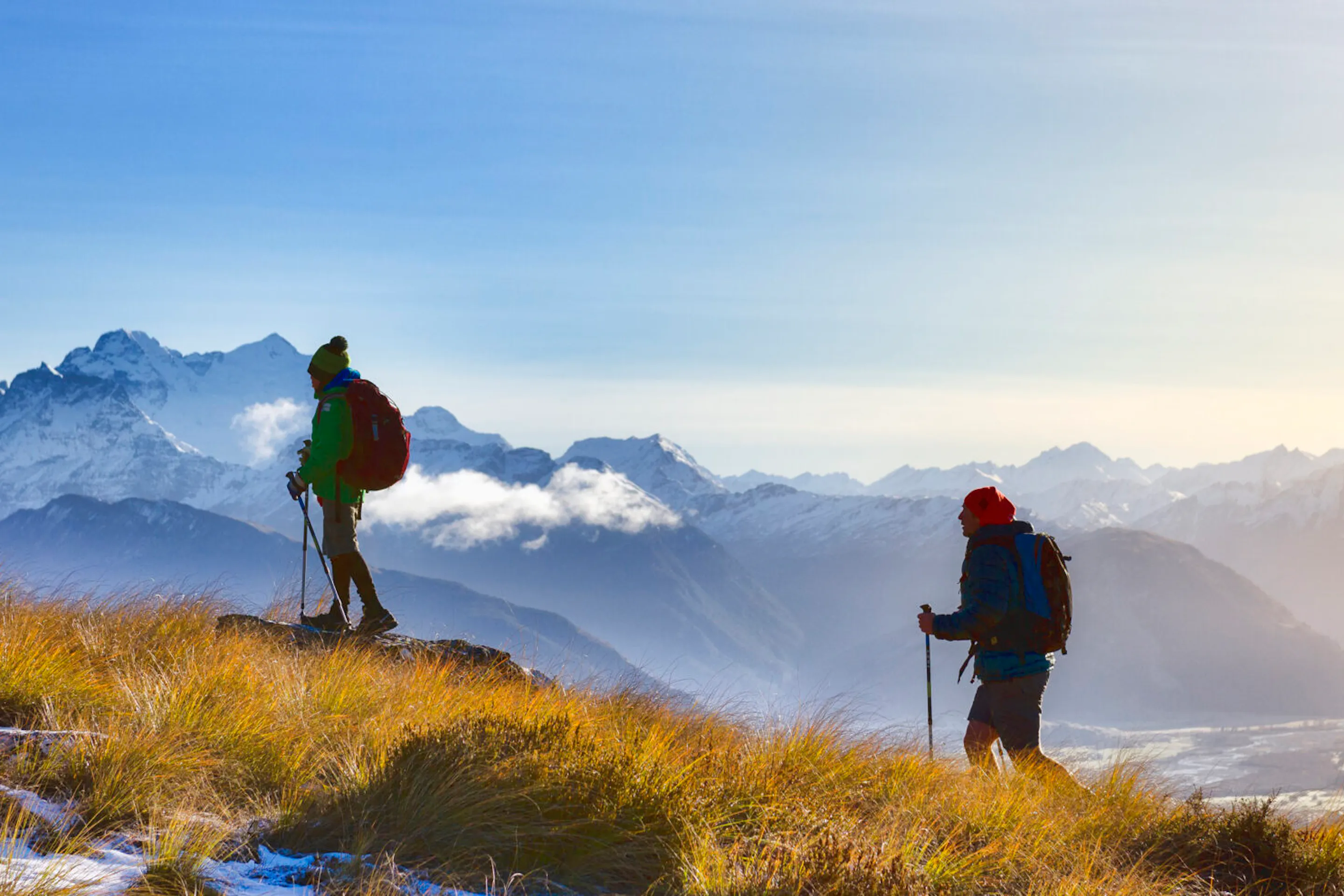 Two hikers stand on a ridge above Fiordland's misty mountains, with layered peaks fading into low cloud below.