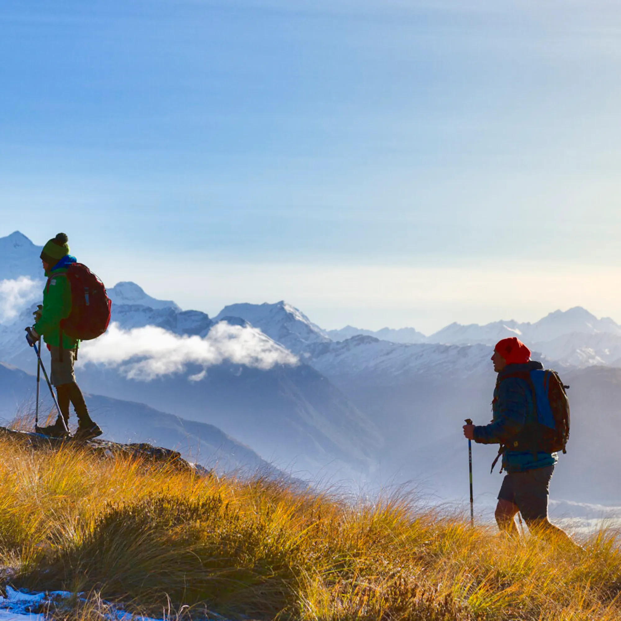 Two hikers stand on a ridge above Fiordland's misty mountains, with layered peaks fading into low cloud below.