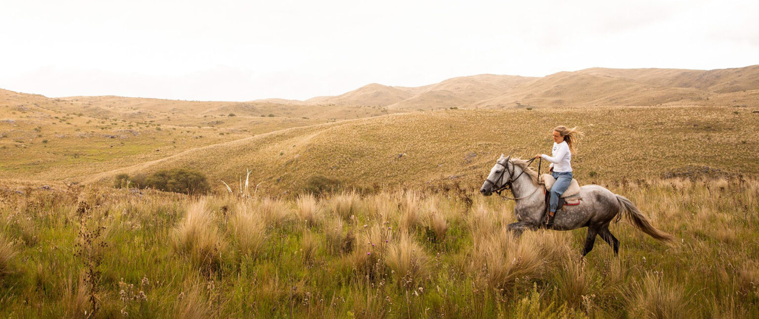 A rider on horseback crossing open grassland near the estancia at Estancia Los Potreros in Cordoba, Argentina.