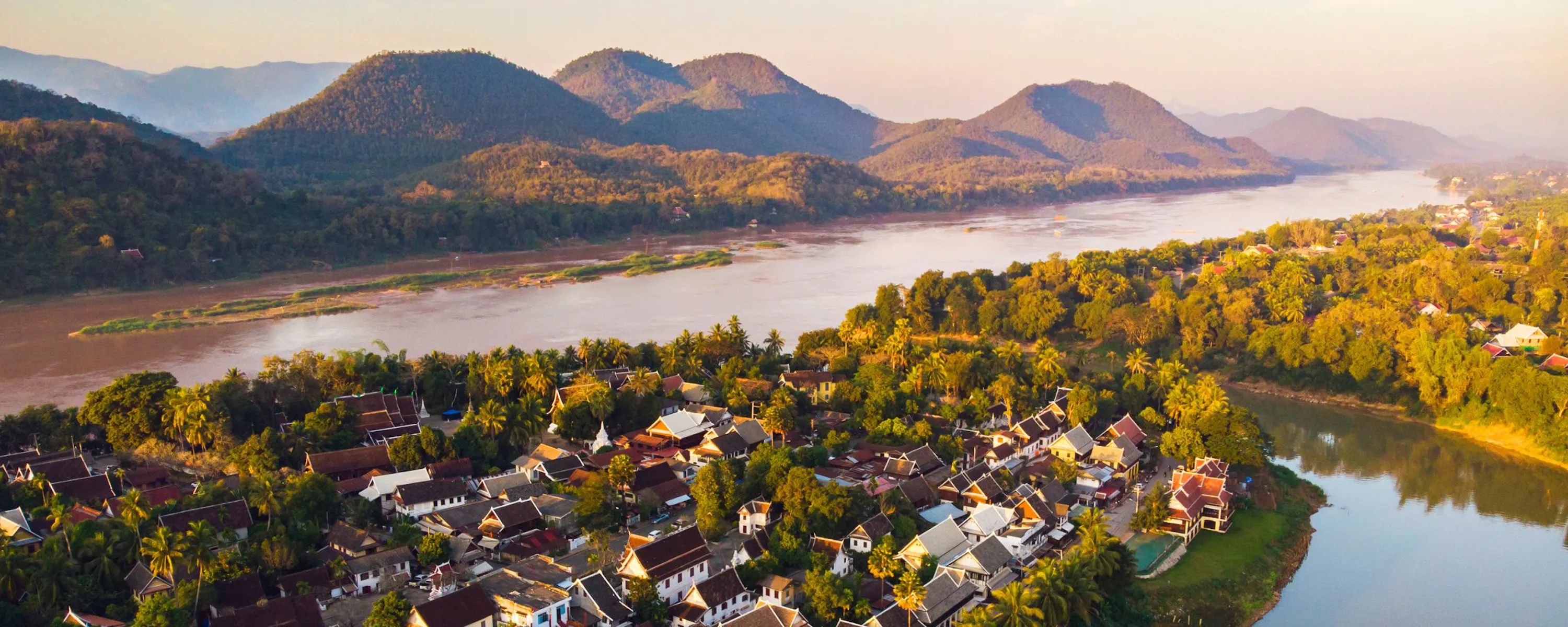 Luang Prabang sits beside a winding river, with sunlit trees, clustered rooftops, and mountains beyond the valley.