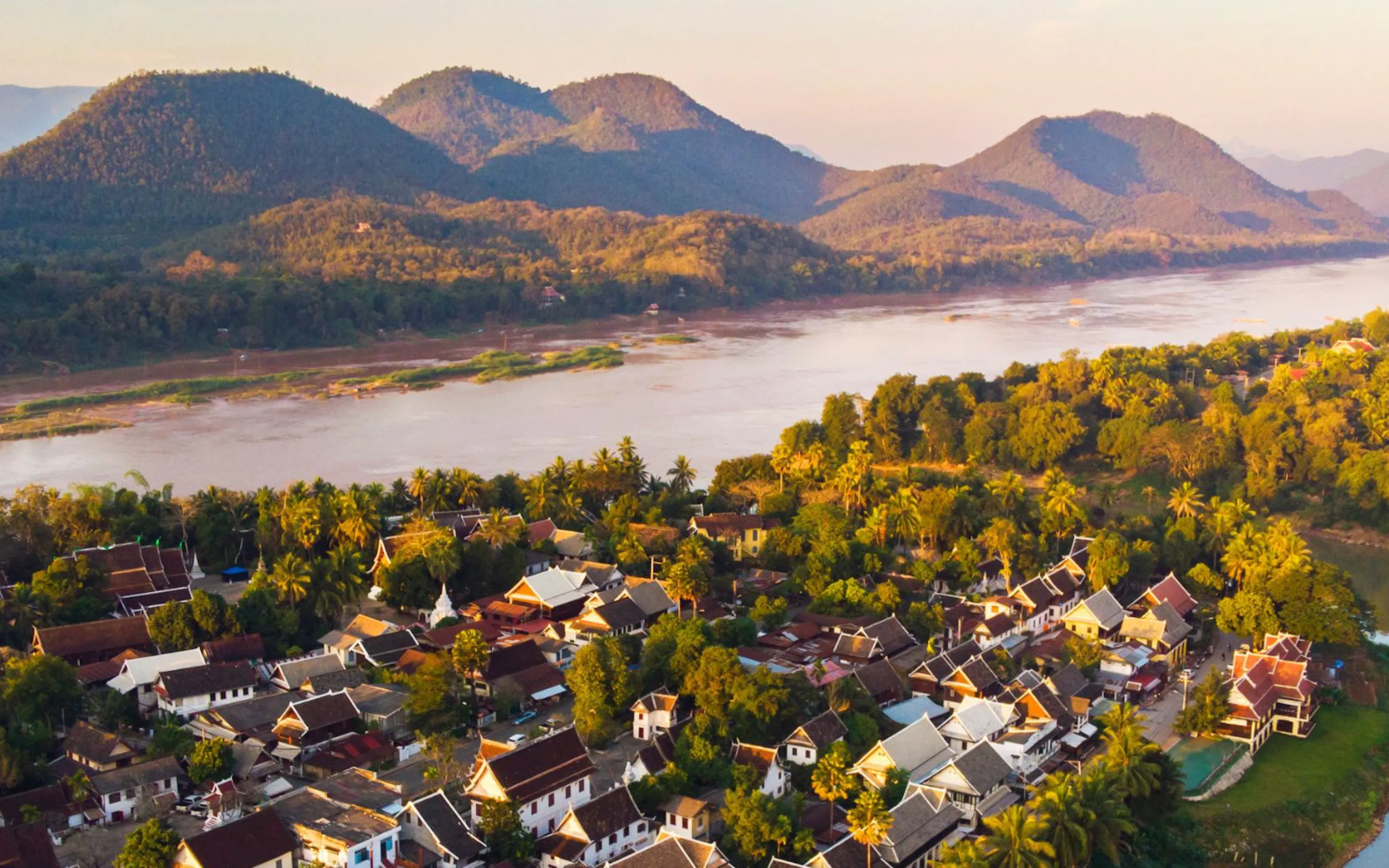 Luang Prabang sits beside a winding river, with sunlit trees, clustered rooftops, and mountains beyond the valley.