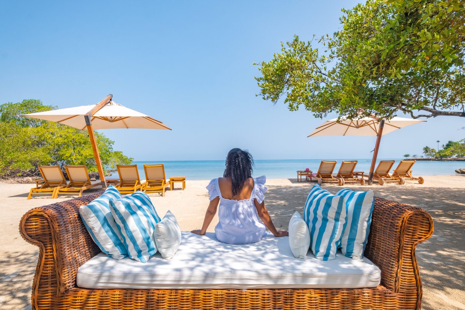 A guest sits on a wicker lounger at Casa San Agustin's beach, facing pale sand, blue water, and umbrellas.
