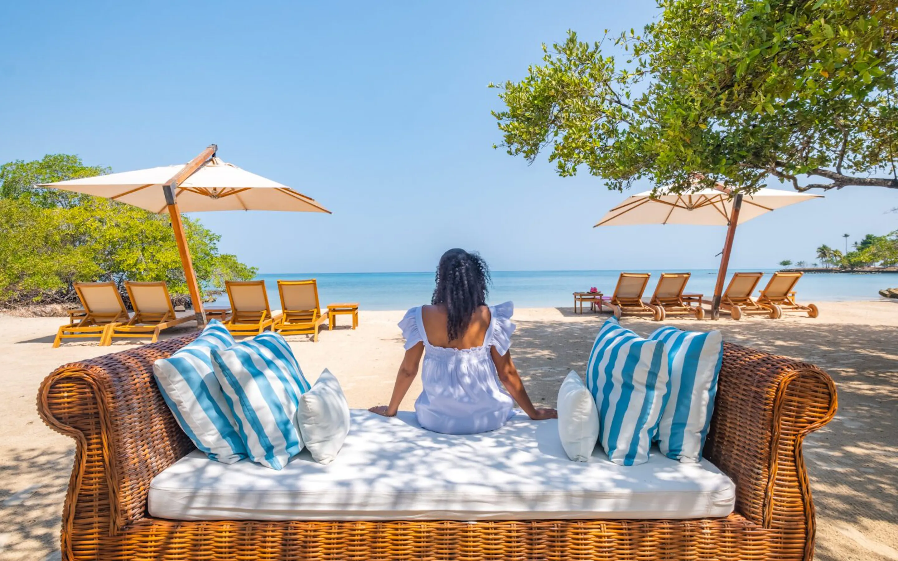 A guest sits on a wicker lounger at Casa San Agustin's beach, facing pale sand, blue water, and umbrellas.