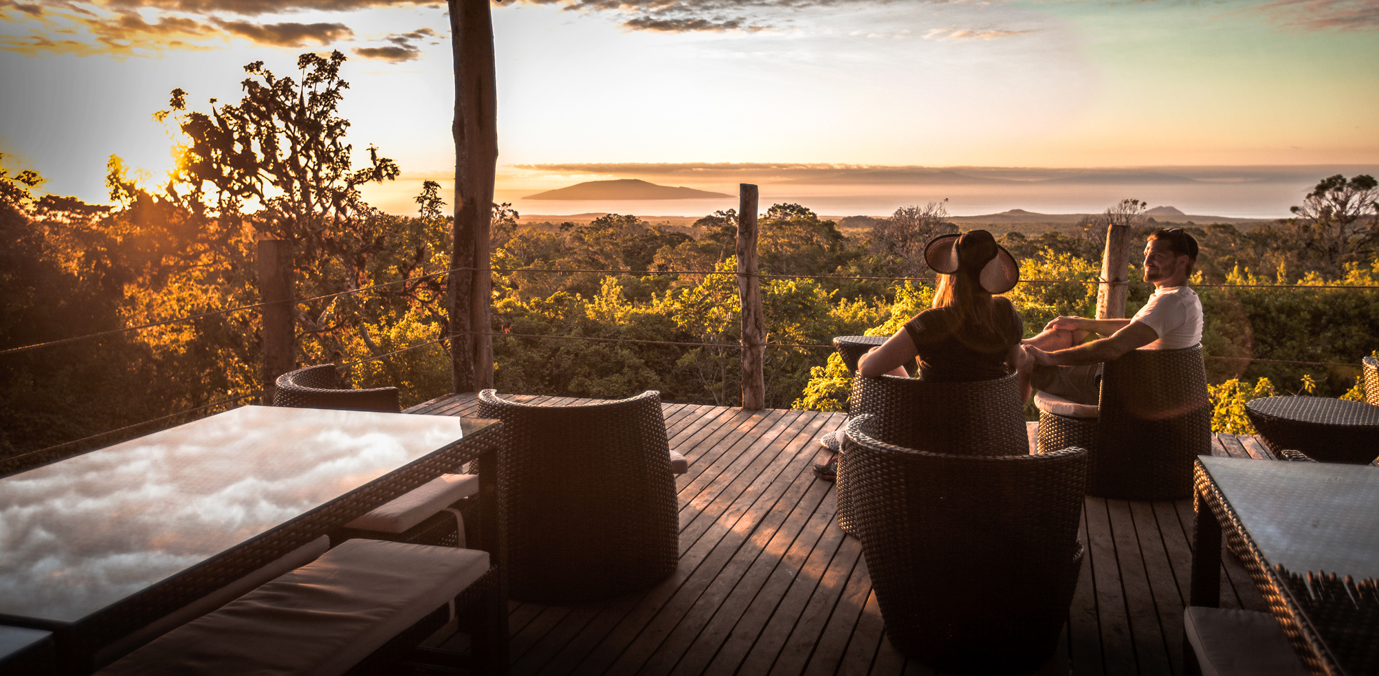A couple relaxes on a deck overlooking the Galapagos, with distant islands and low water below the viewpoint.