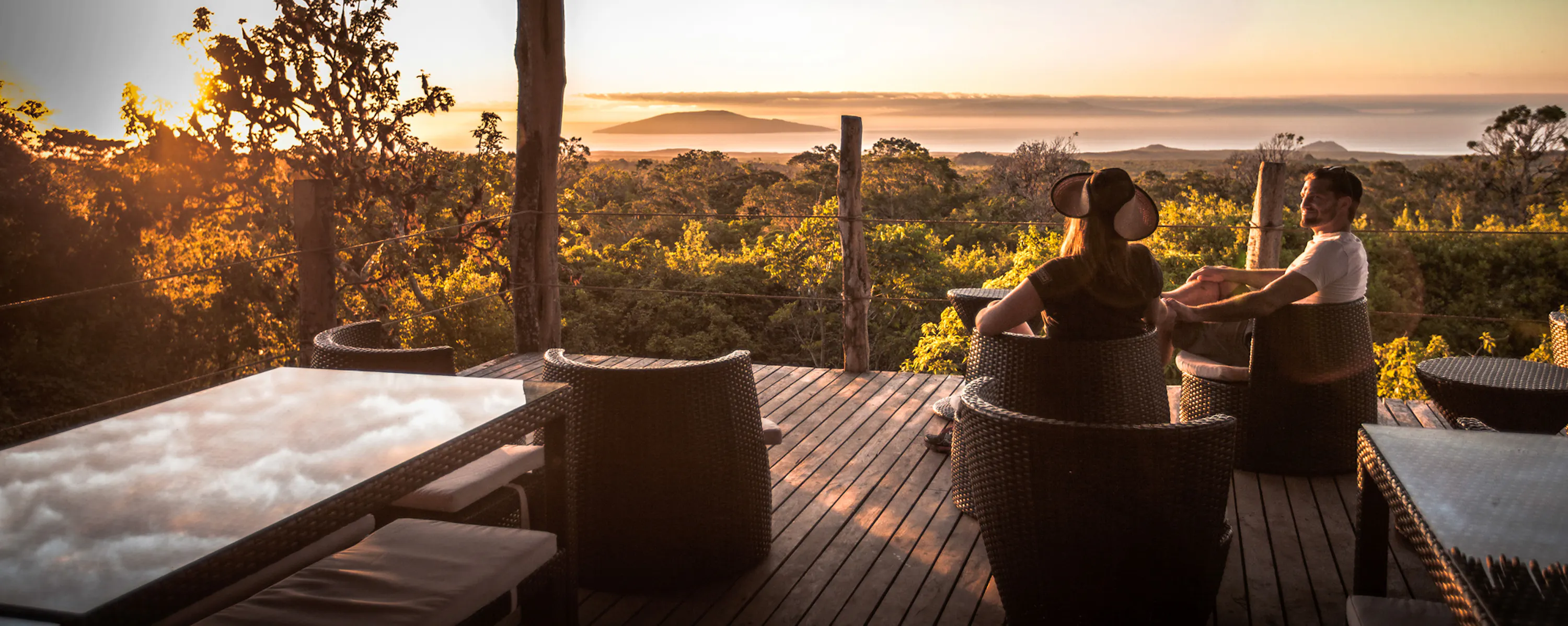 A couple relaxes on a deck overlooking the Galapagos, with distant islands and low water below the viewpoint.