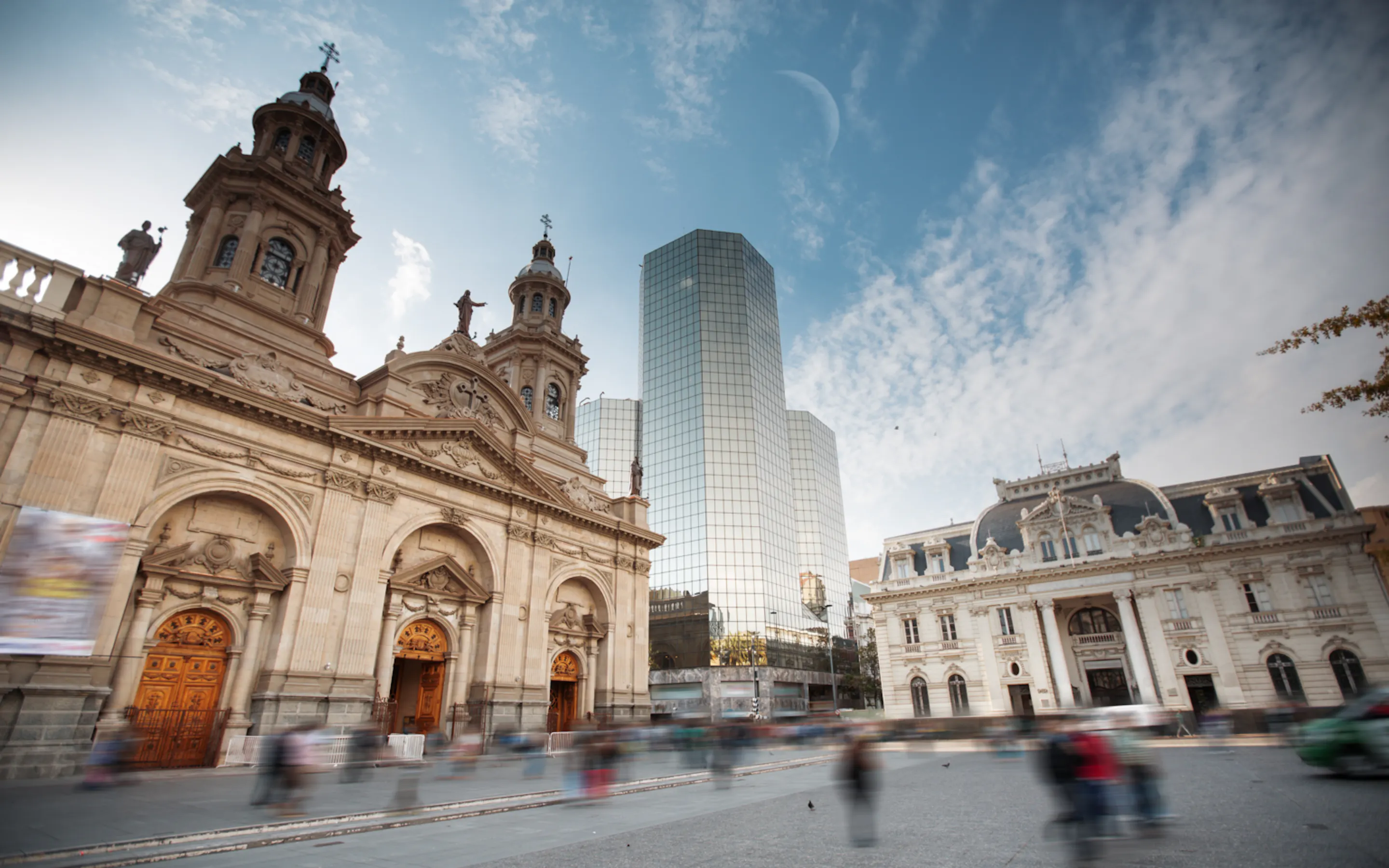 Historic buildings and glass towers rise in central Santiago, with a partly cloudy sky and layered city streets below.