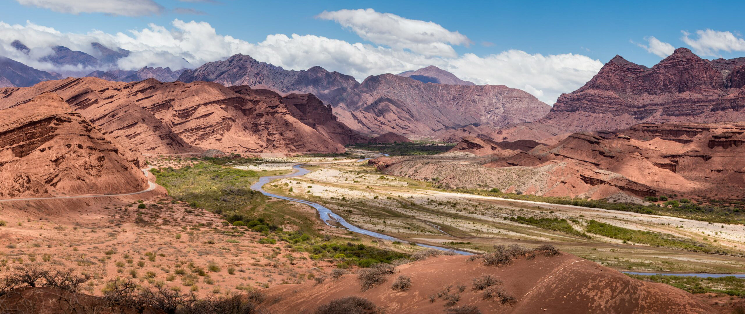 A winding river runs through red rock valleys and layered mountains in northwest Argentina beneath bright clouds.