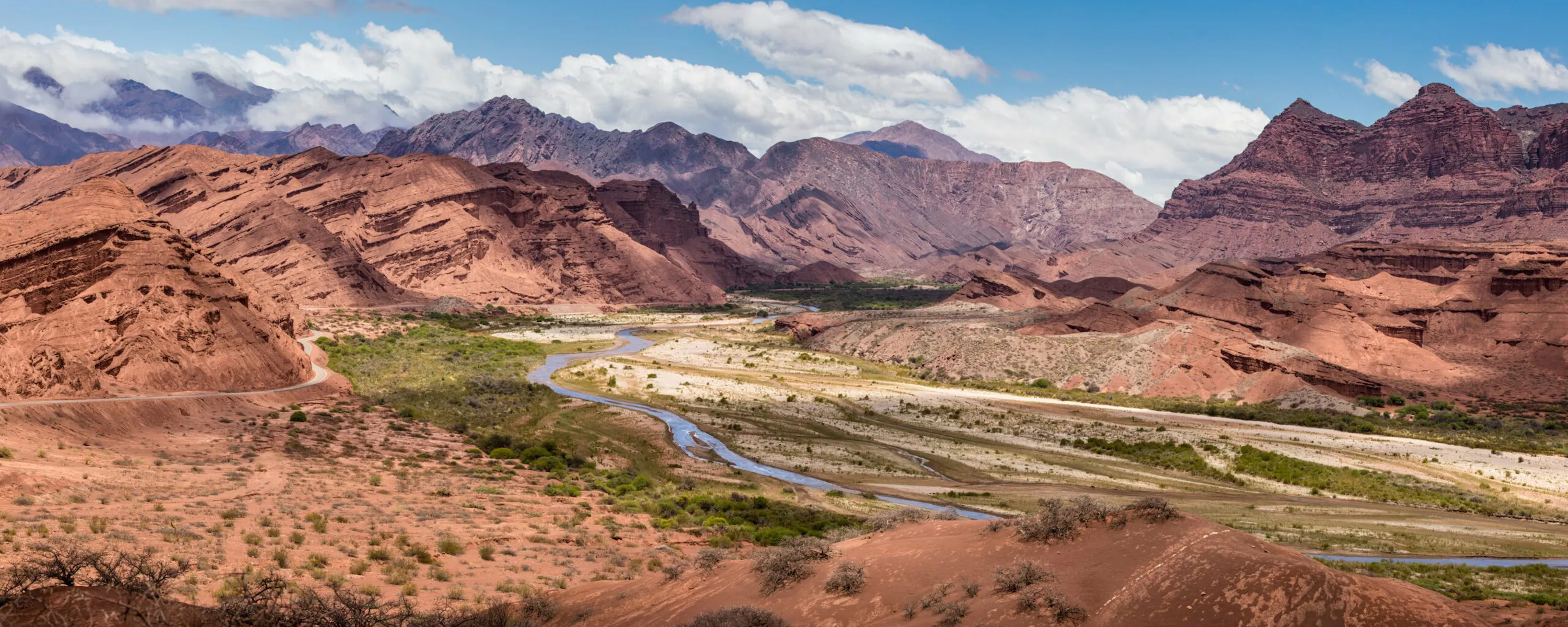 A winding river runs through red rock valleys and layered mountains in northwest Argentina beneath bright clouds.